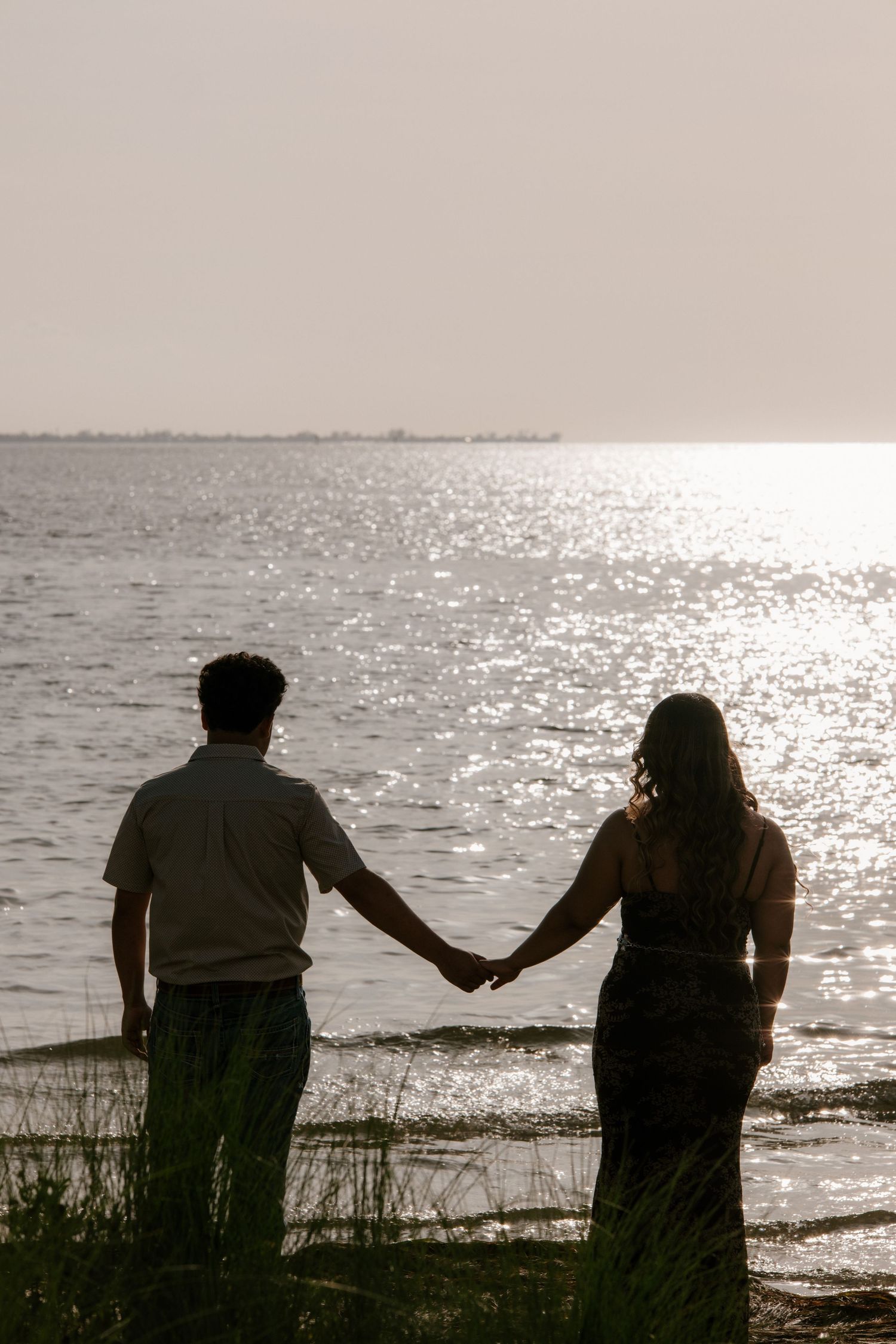 Silhouette of a couple holding hands facing a sparkling ocean at sunset near a grassy shoreline.
