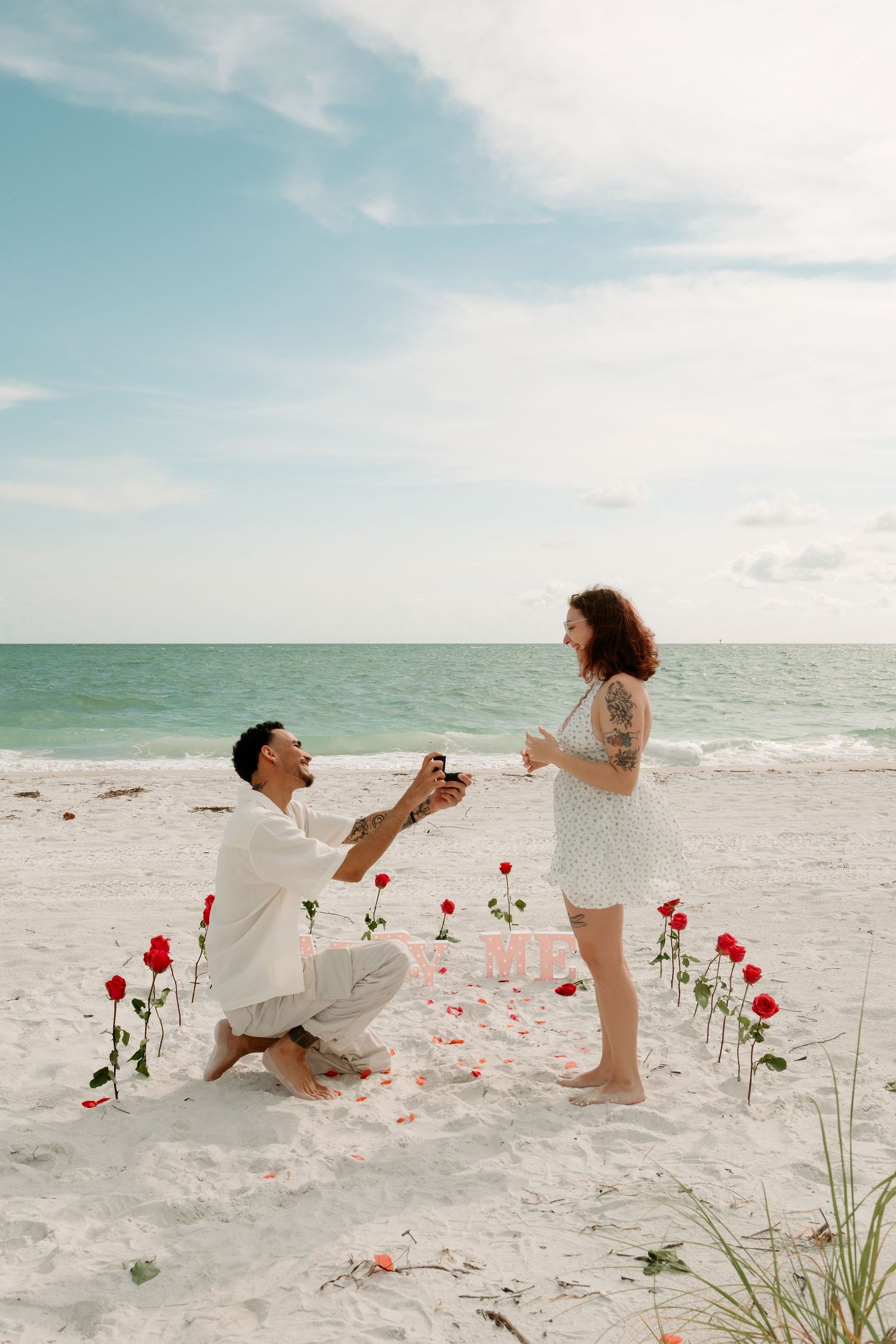 Man proposes on bended knee with red roses on a sandy beach, as woman reacts with joy by the ocean.