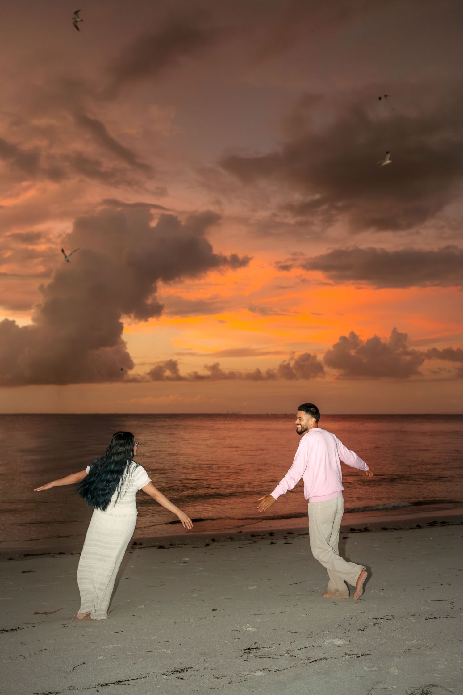 Couple dancing barefoot on a beach at sunset with dramatic orange clouds and ocean waves in the background.