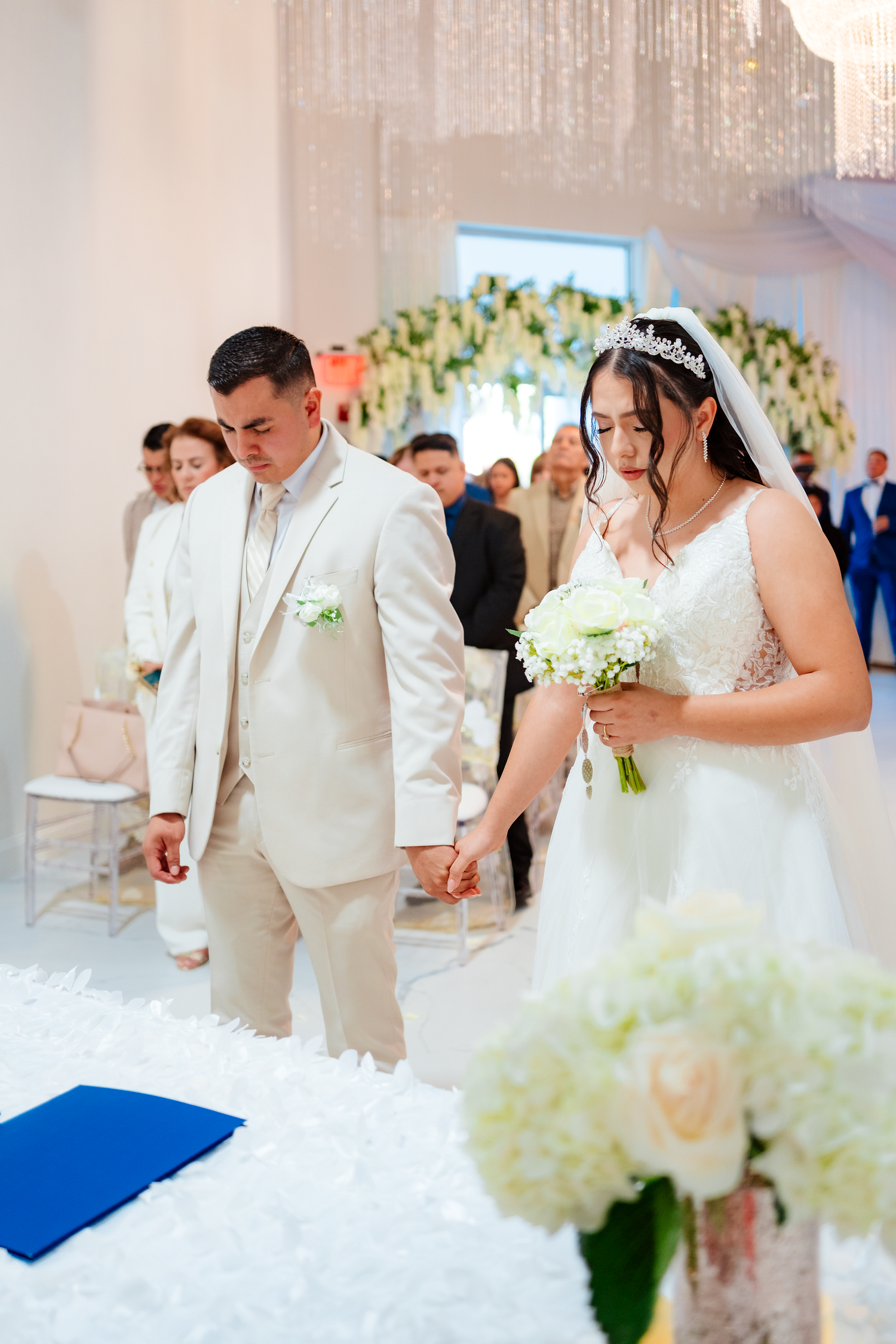 Bride and groom holding hands at wedding ceremony altar, bride wearing tiara and veil holding white bouquet.