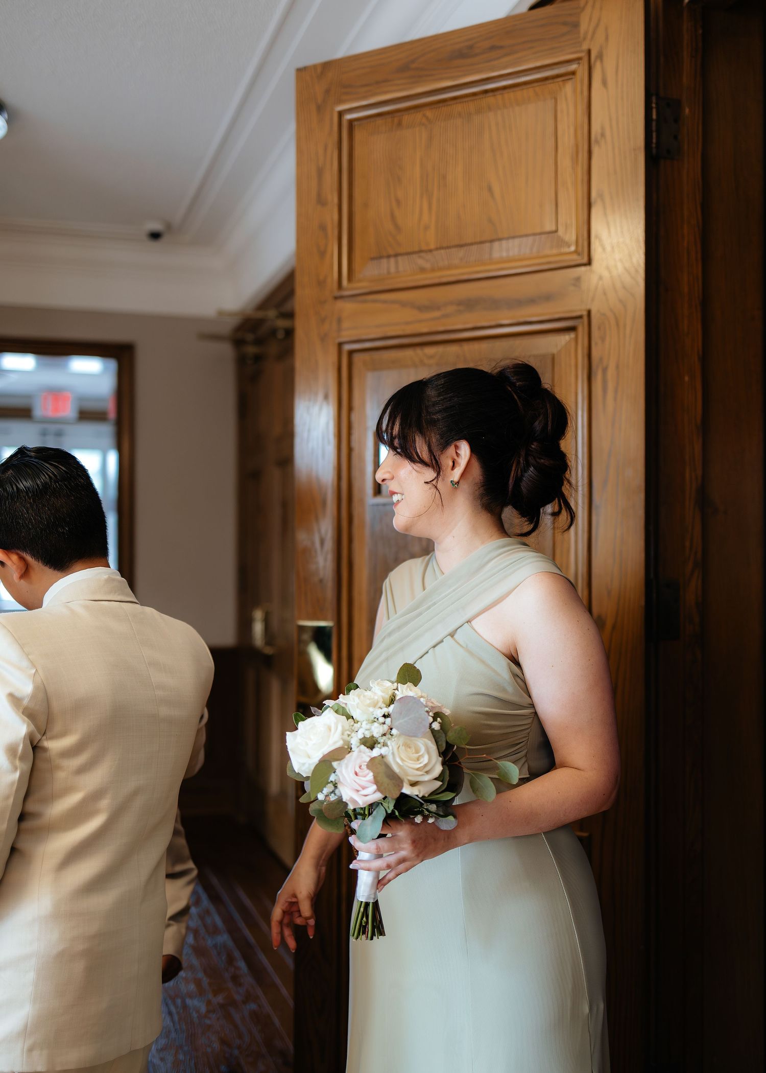 Elegant bridesmaid in sage green dress holds white rose bouquet near wooden door before wedding.