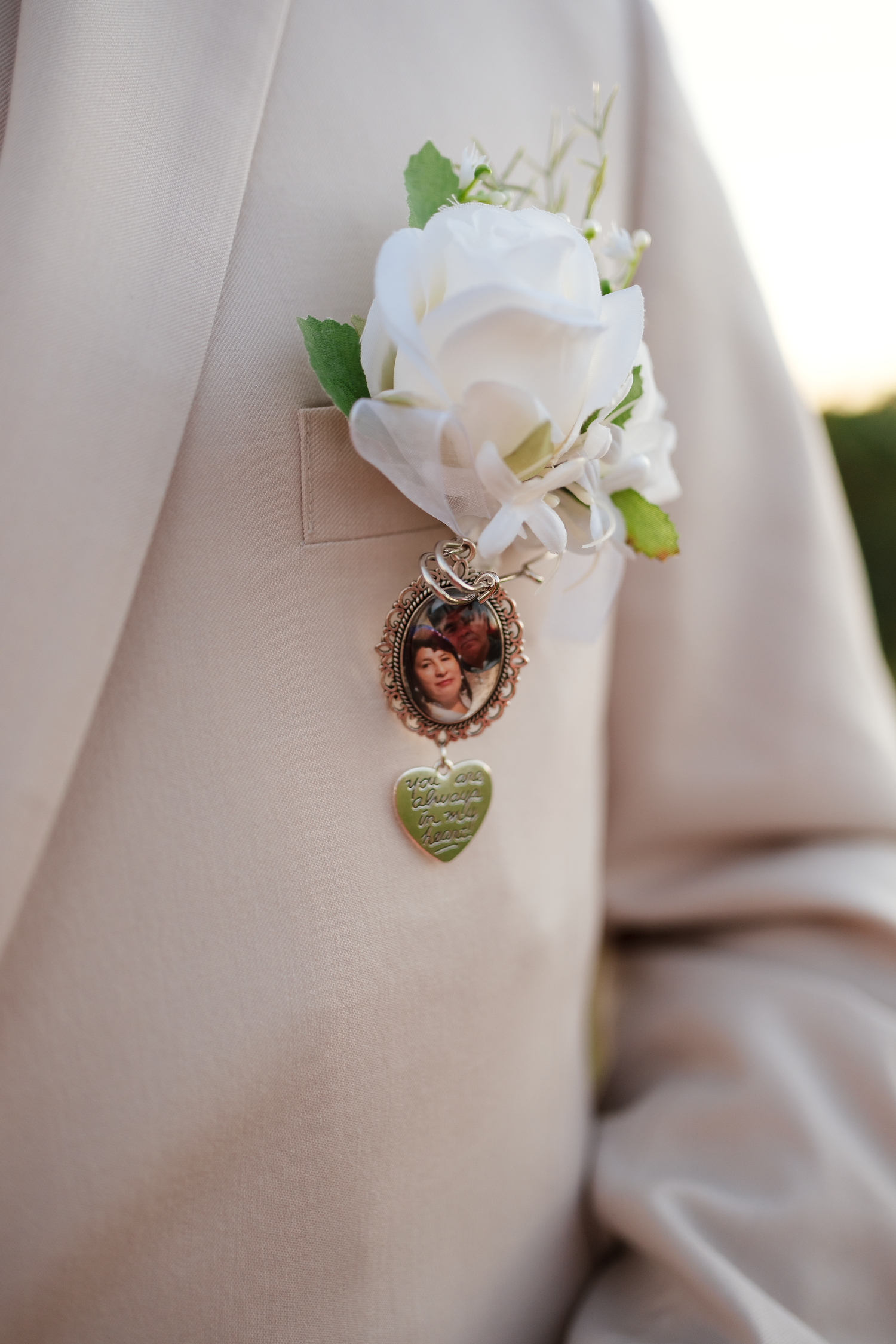 Close-up of groom's boutonniere with white rose and memorial photo locket charm on lapel.