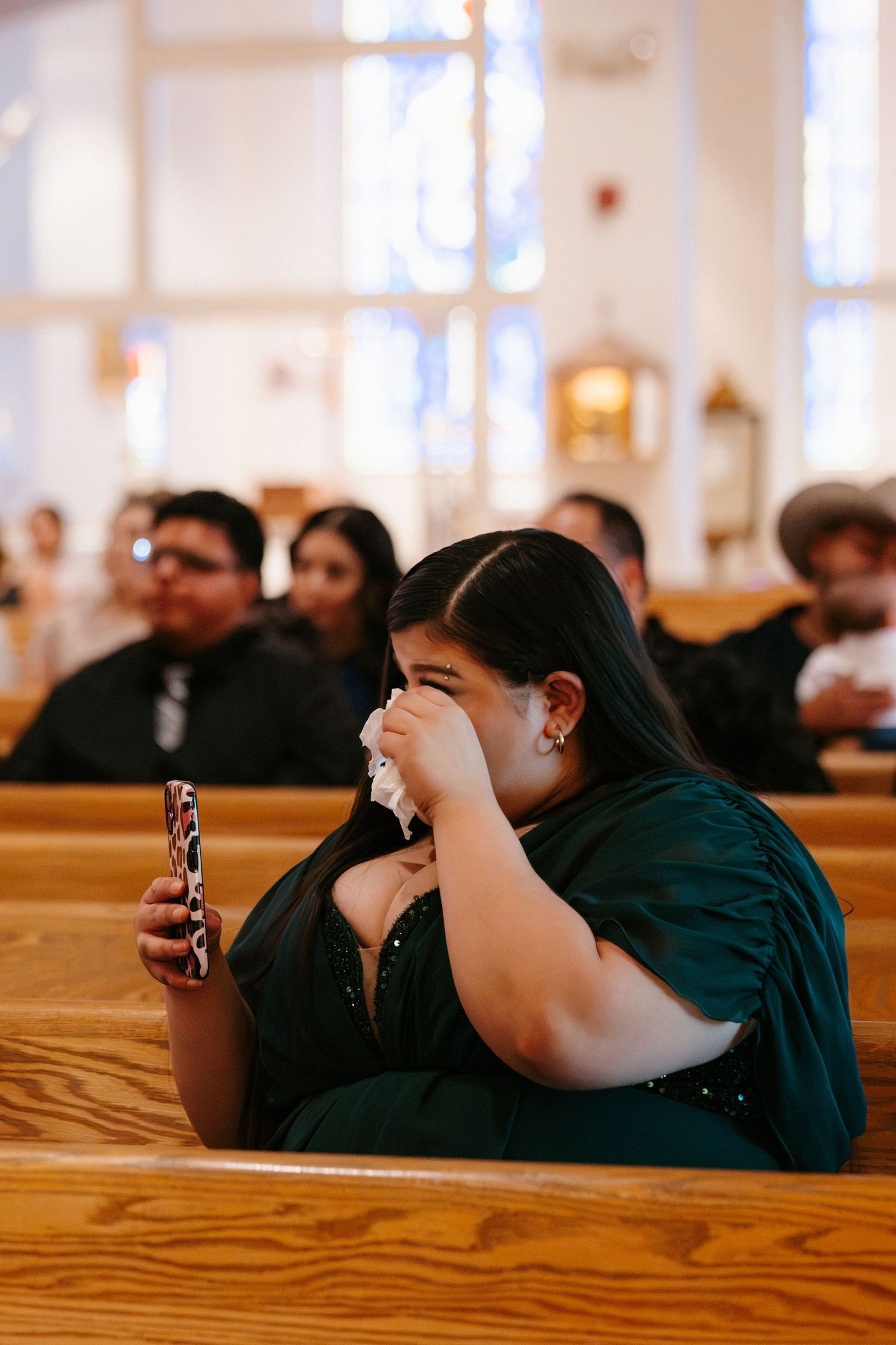 A woman in a green dress wipes tears with a tissue while holding a phone in a church pew.