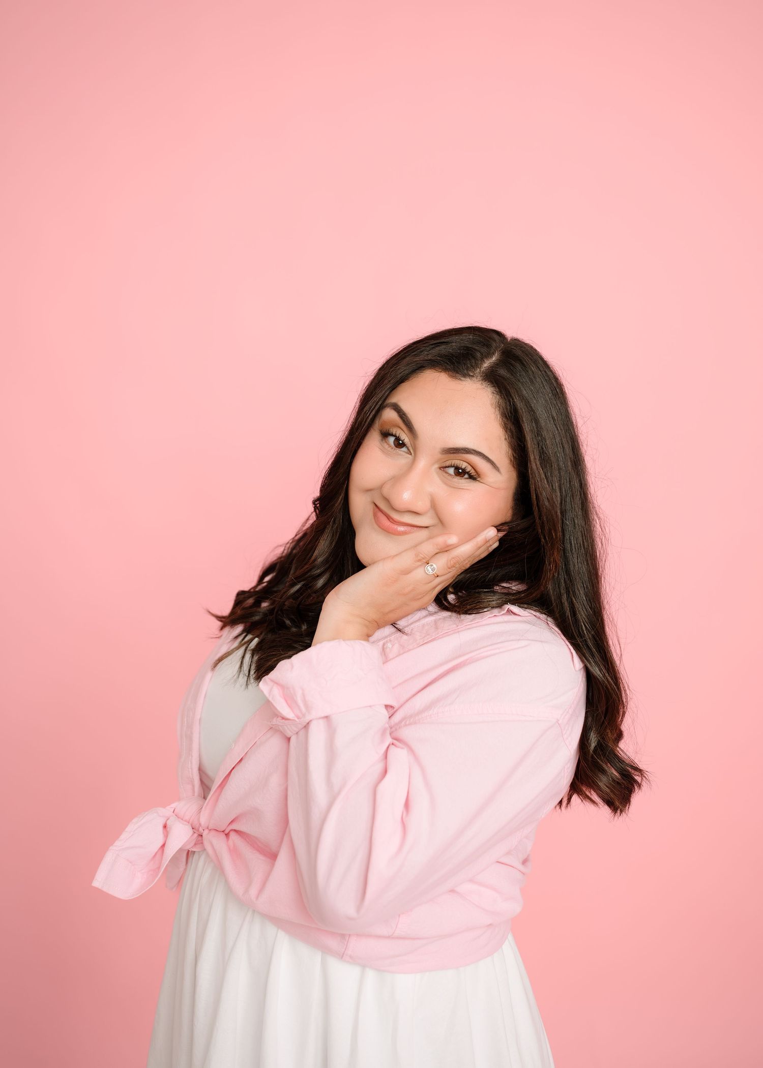 Young woman in pink tie-front top poses playfully against pink background with hand on cheek.