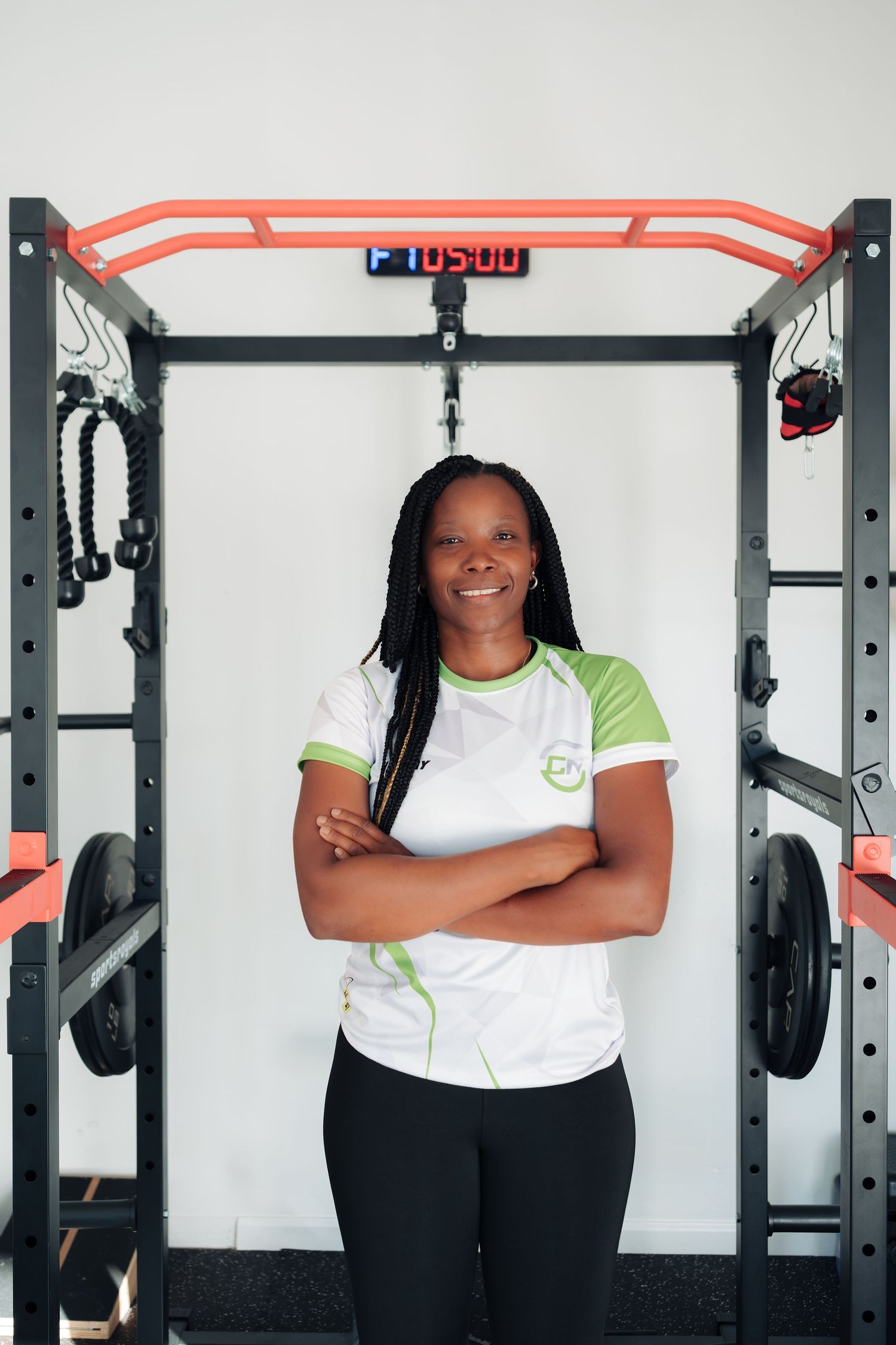 Smiling female fitness trainer in white and green shirt stands arms crossed in front of gym power rack equipment.