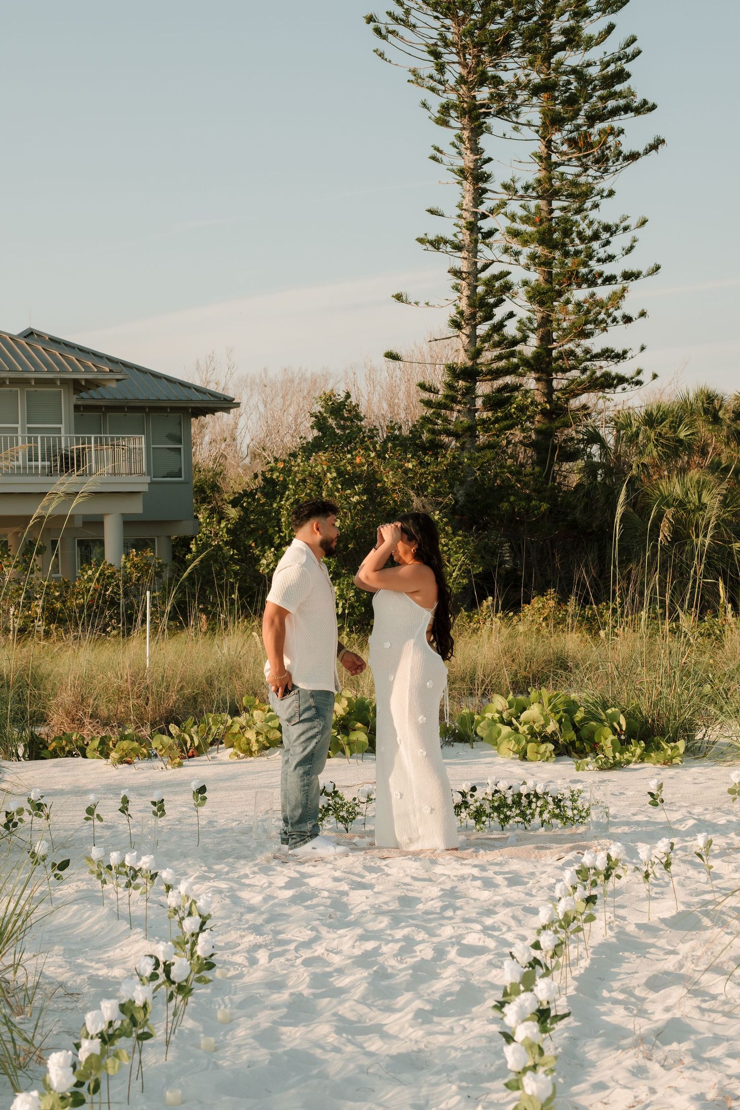 Couple in white attire sharing a romantic moment on a sandy beach with floral arrangements and tall pine trees at sunset.