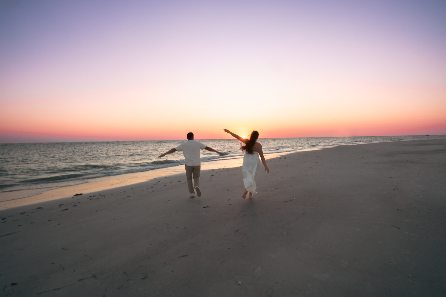 Couple holding hands running on a sandy beach at sunset with colorful pink and purple sky over calm ocean waters.