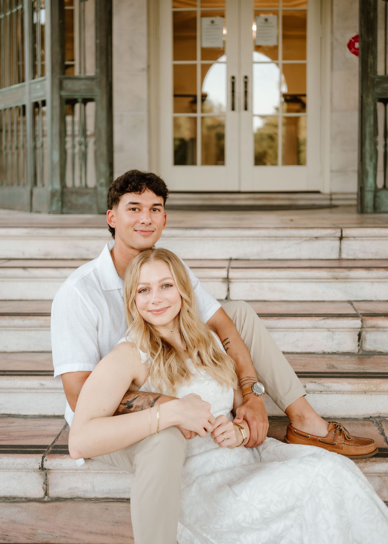A couple sits on marble steps outside elegant building, woman in white dress leaning on man in white shirt.