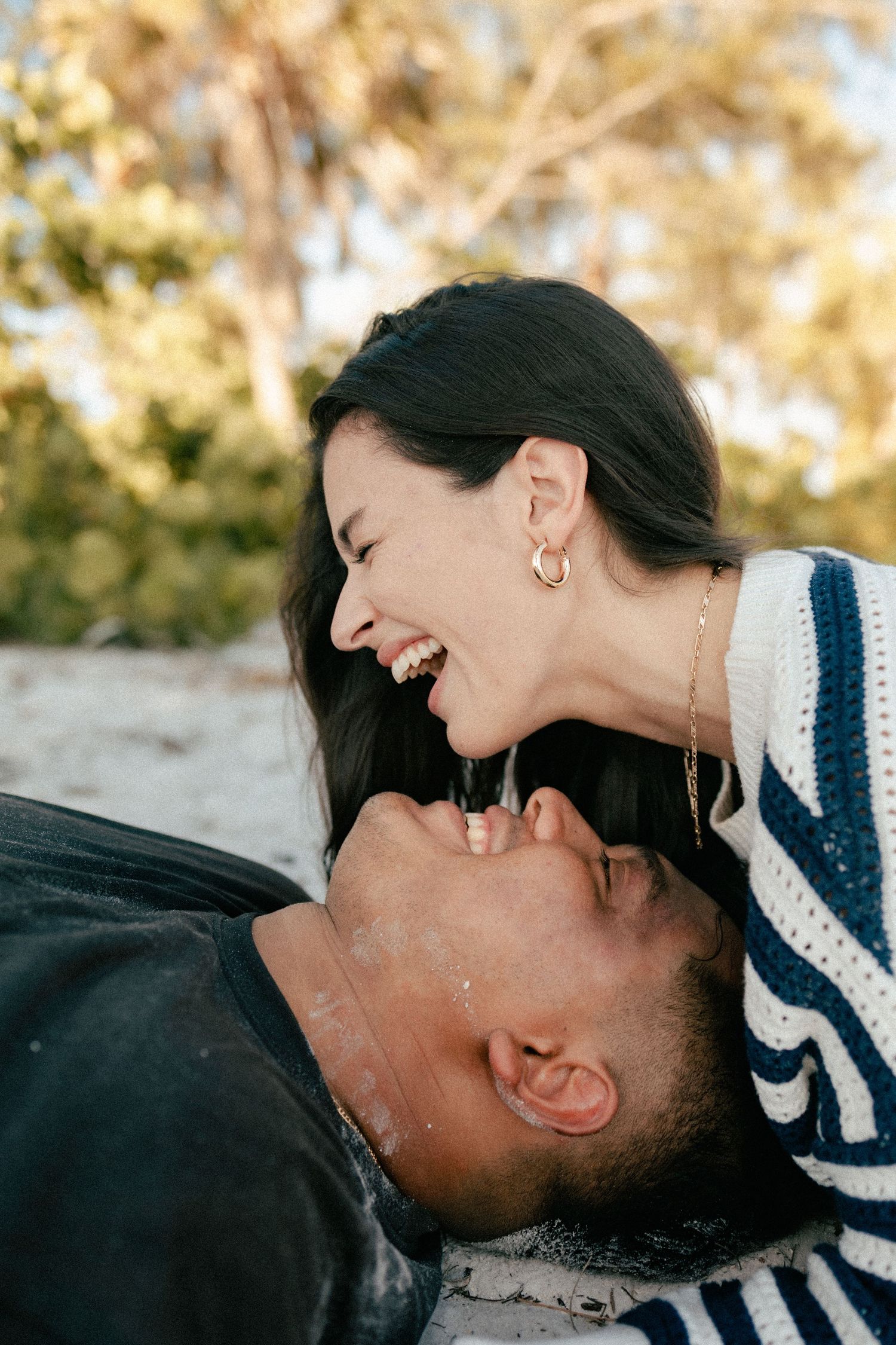 A laughing couple sharing a joyful moment outdoors, woman leaning over smiling man lying back, trees in background.