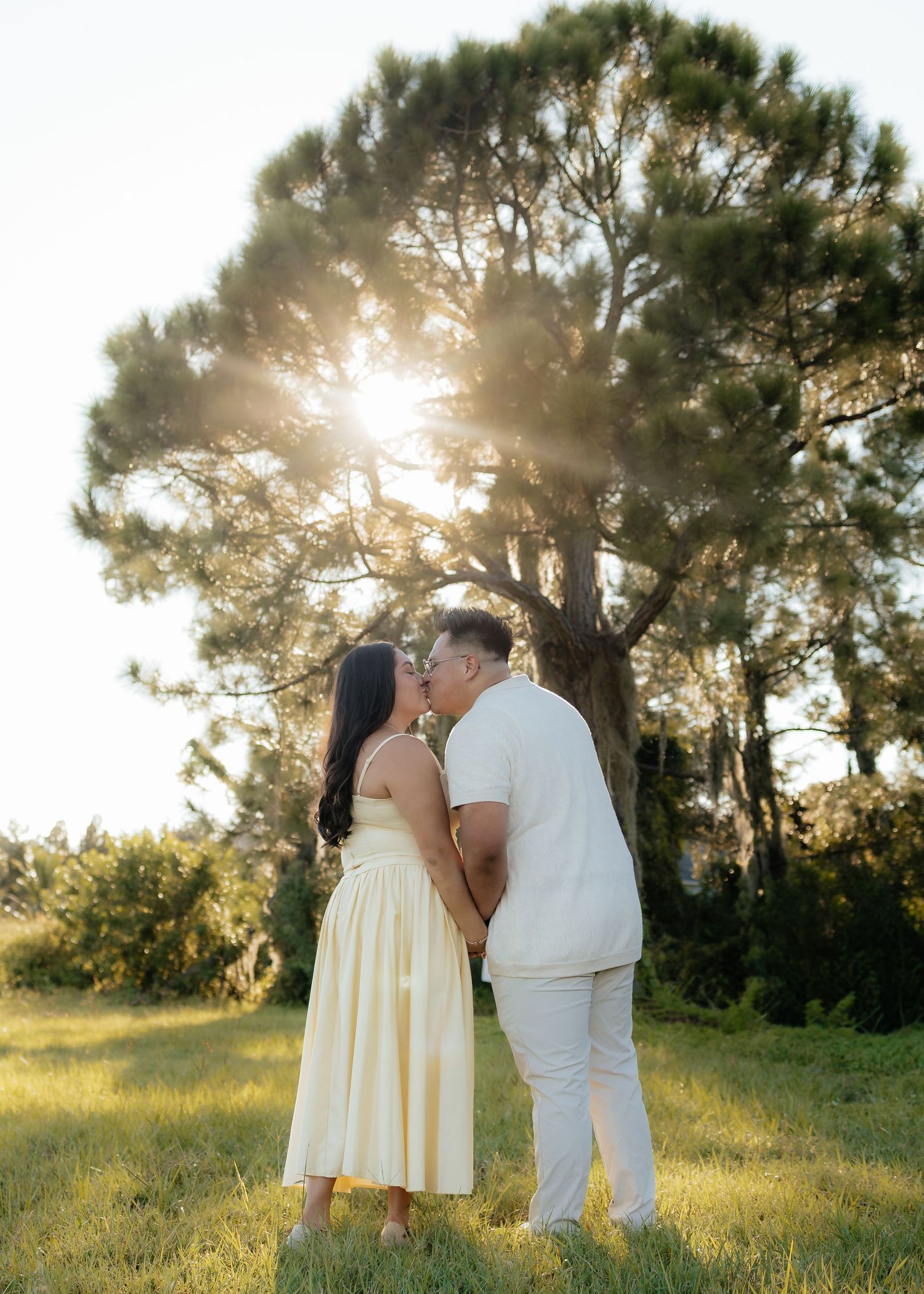Couple sharing a romantic kiss in a sunlit park, woman in yellow dress and man in white outfit, with a large tree backdrop.