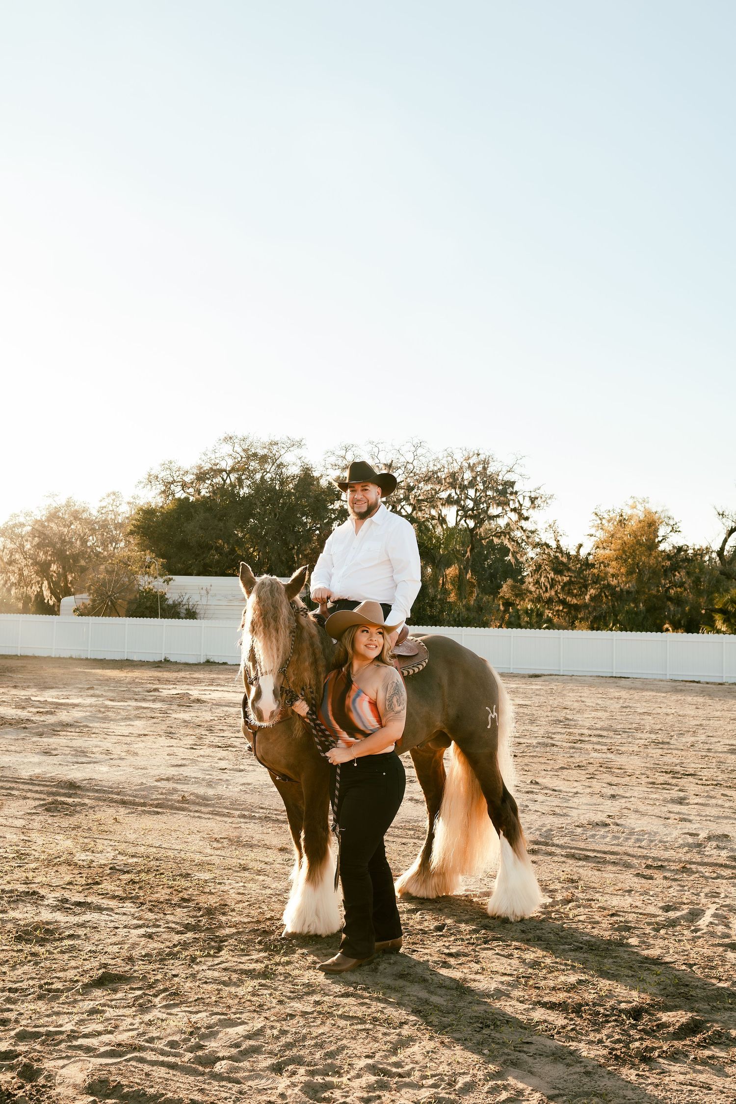 A man in a cowboy hat sits atop a brown horse while a woman in a cowboy hat stands beside it in a sandy arena.
