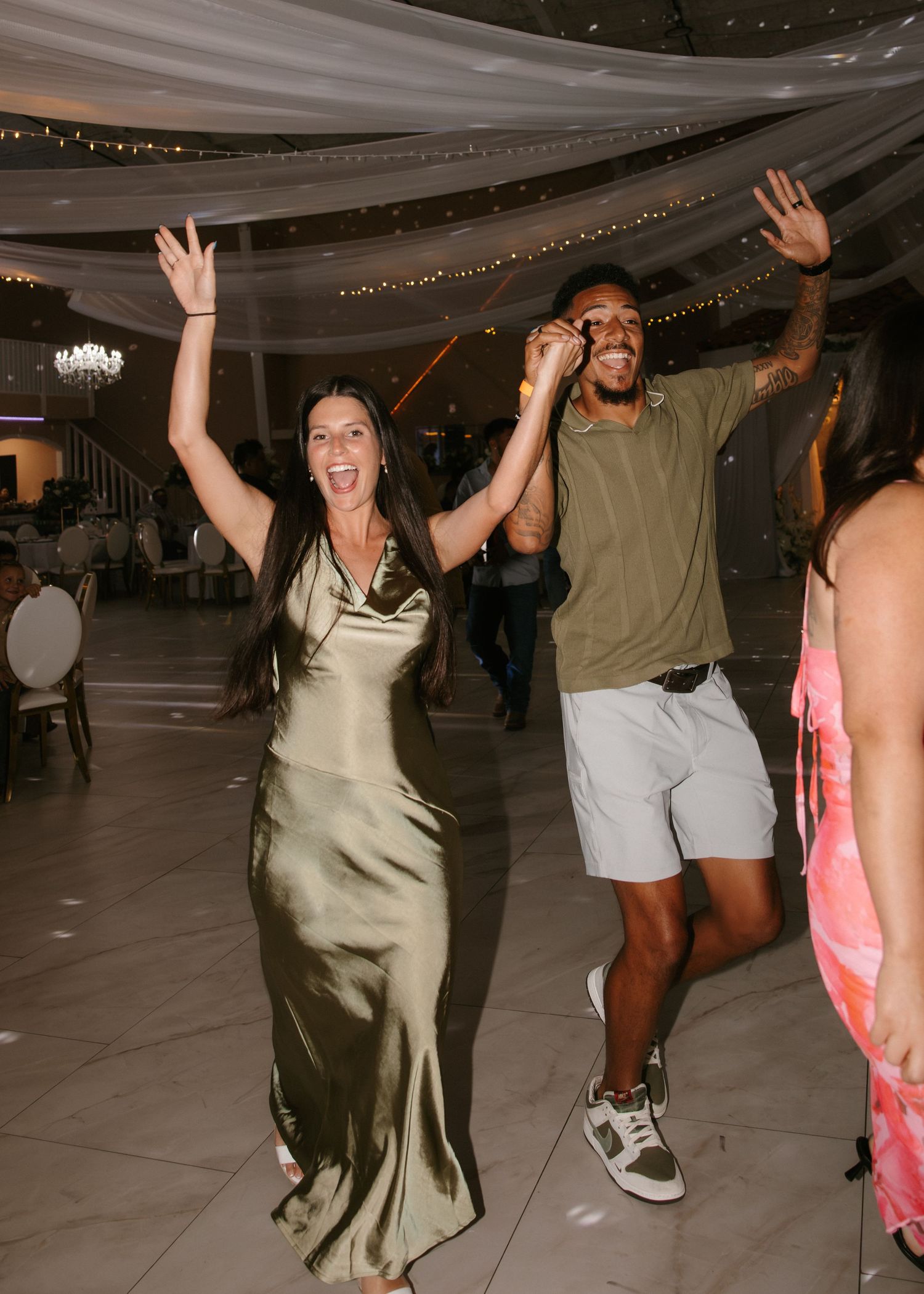Guests in elegant attire celebrate joyfully on wedding reception dance floor with raised hands.