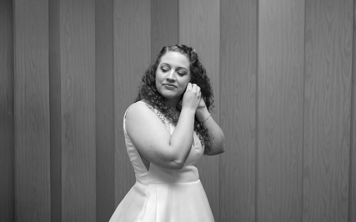 Black and white portrait of a young woman in a white dress posing gracefully against a wooden panel wall.