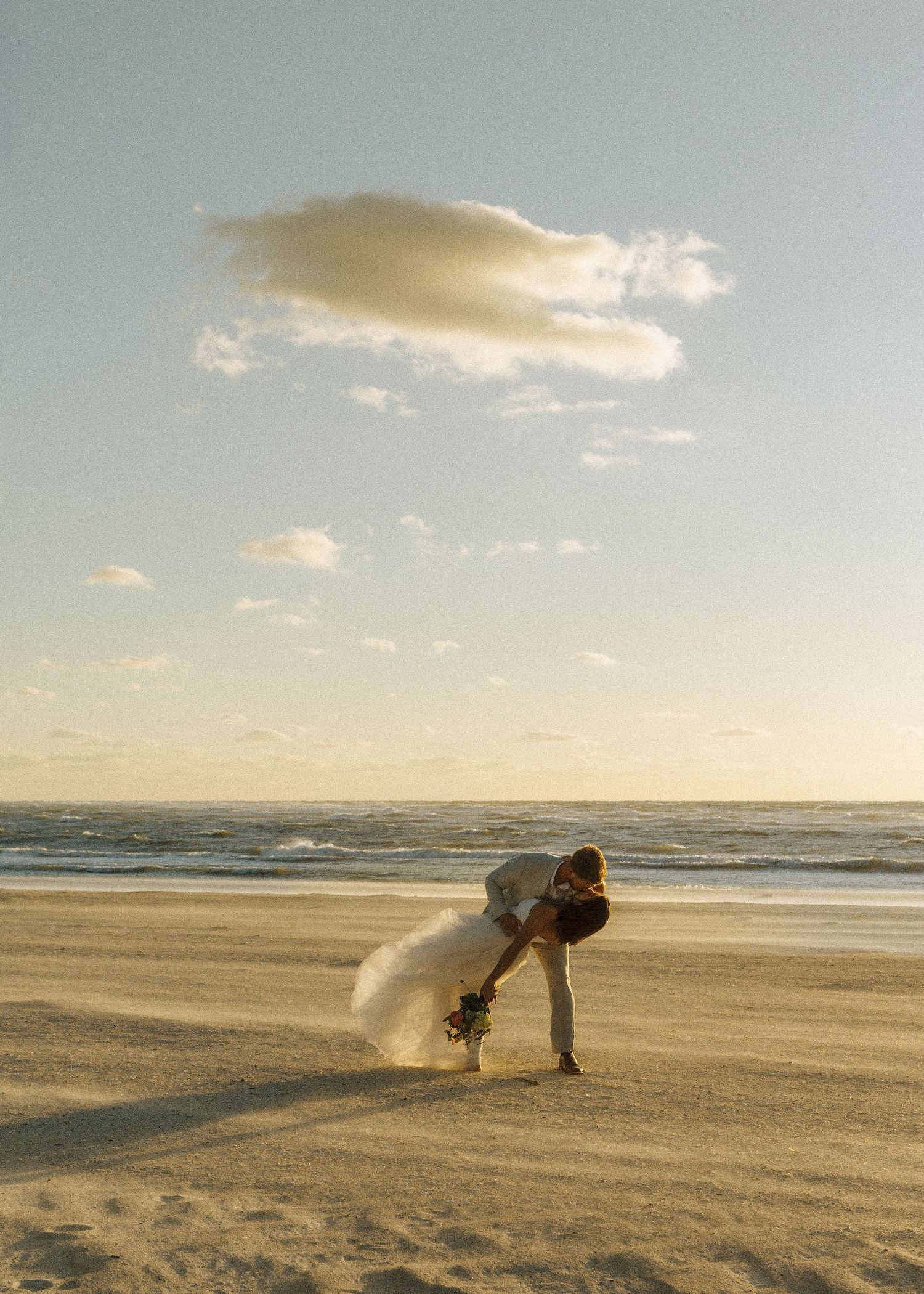Couple sharing a romantic dip kiss on a sandy beach at sunset, bride in white gown holding bouquet.