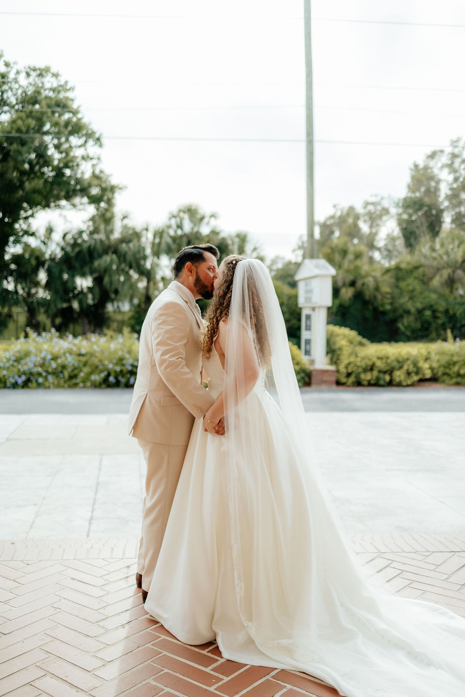 Bride and groom share a romantic kiss on a brick pathway surrounded by lush garden greenery.