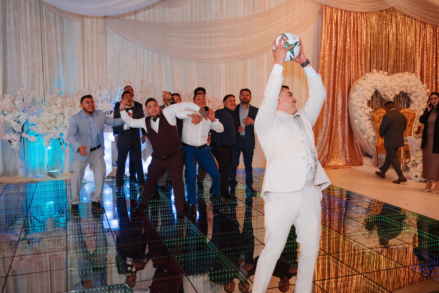 Groom catching bouquet toss surrounded by groomsmen on mirrored wedding reception dance floor.