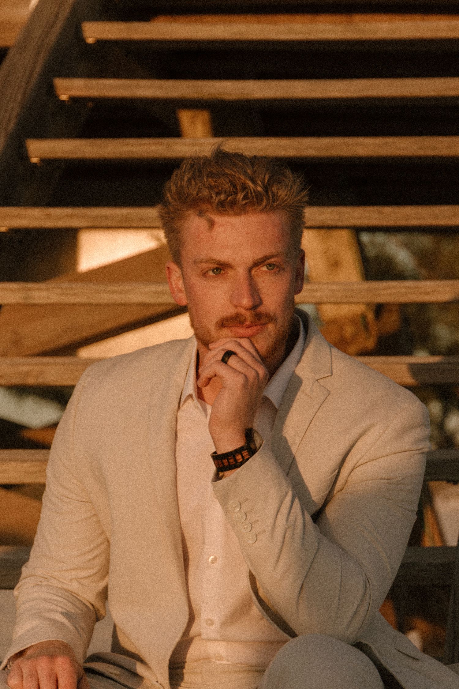 Young blond man in beige suit sitting on wooden steps, resting chin on hand in thoughtful pose.