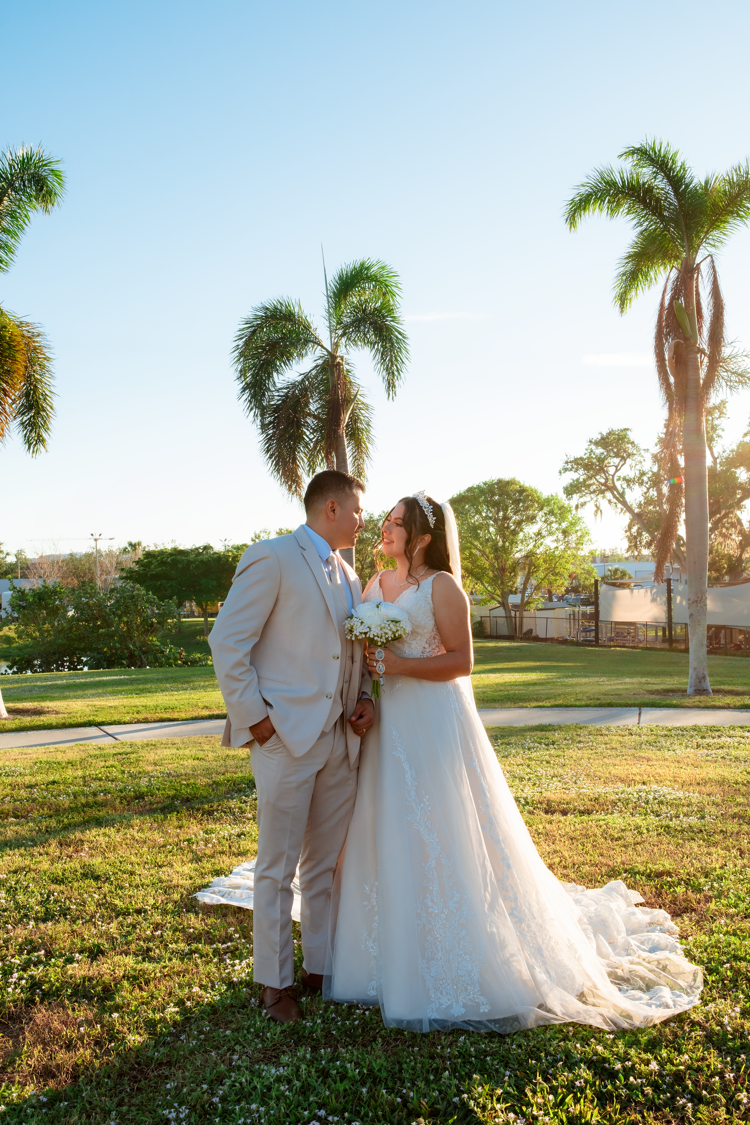 Bride and groom share a romantic moment surrounded by tall palm trees on a sunny wedding day.