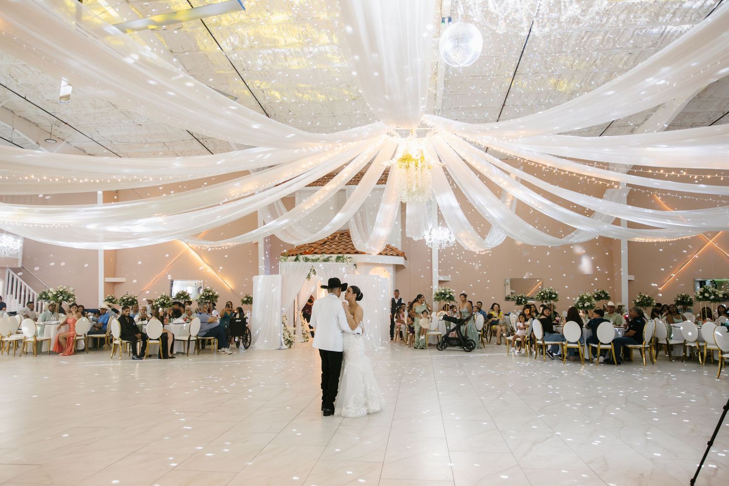 Bride and groom share first dance under elegant white draped ceiling in a grand ballroom wedding reception.