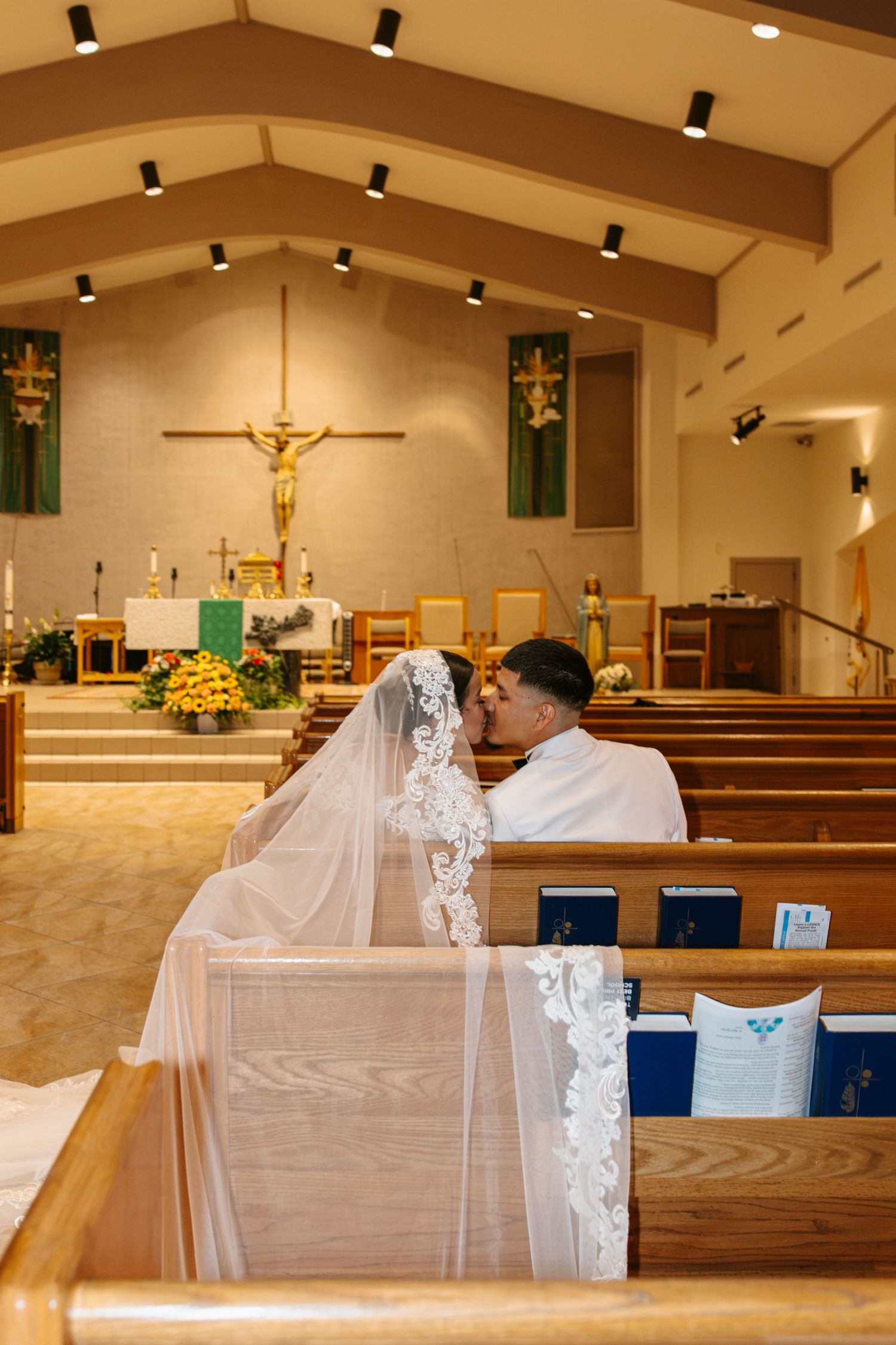 Bride and groom sitting together in church pews during wedding ceremony with altar and crucifix in background.