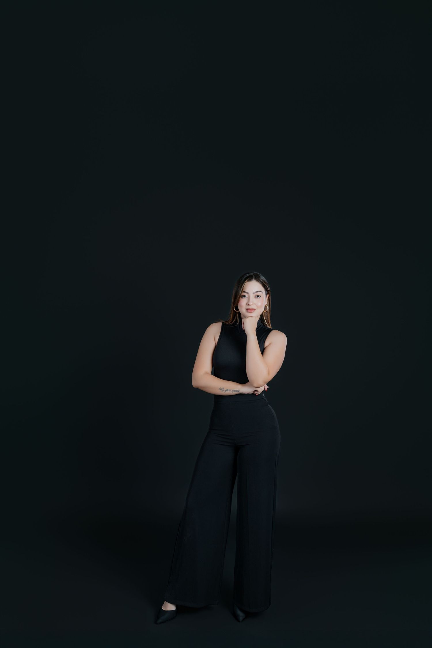 Woman in black sleeveless top and wide-leg pants posing thoughtfully against dark background.