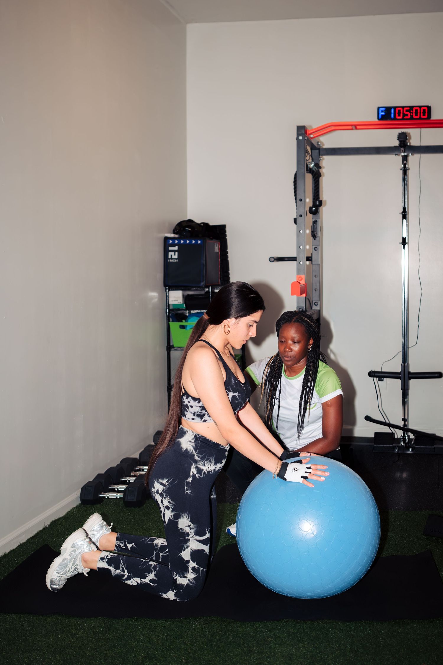 Two women working out with a blue stability ball in a home gym with rubber flooring and workout equipment.