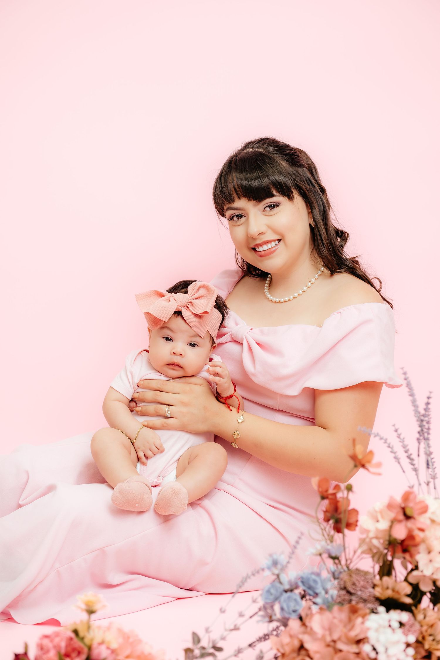 A smiling woman in a pink dress holds her baby girl, both dressed in pink, posed with flowers on a pink background.