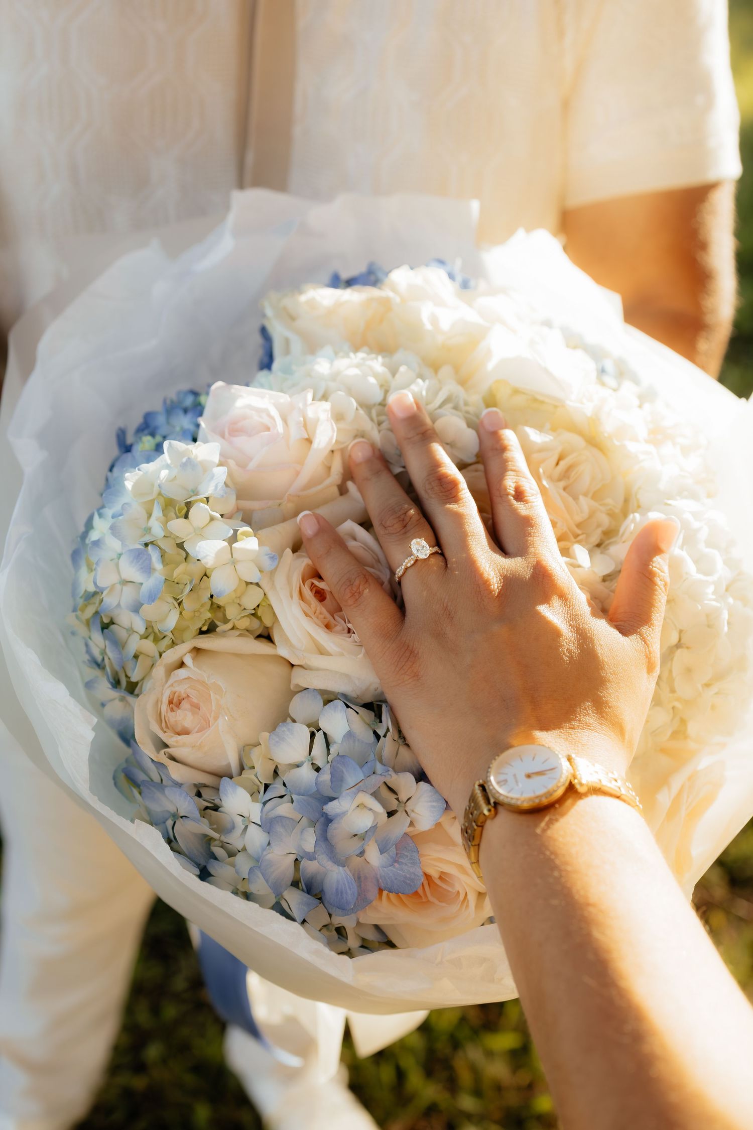 Couple displaying engagement and wedding rings on a bridal bouquet of white roses and blue hydrangeas.