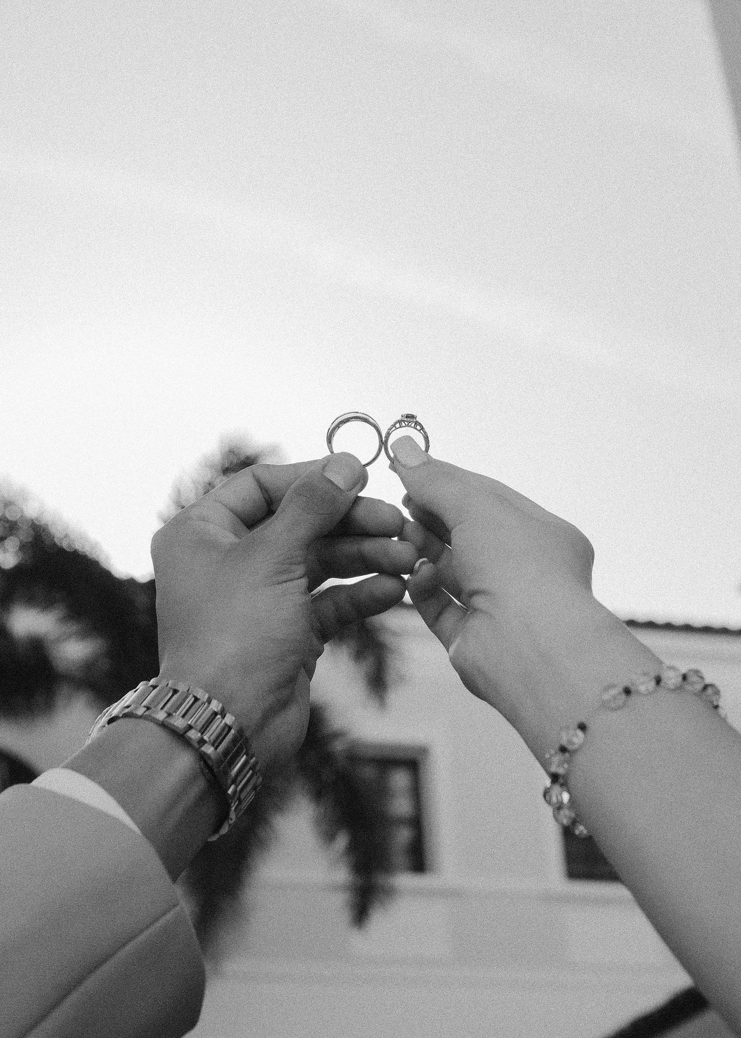 Black and white photo of two hands holding wedding rings up toward the sky, one wearing a watch and one a bracelet.