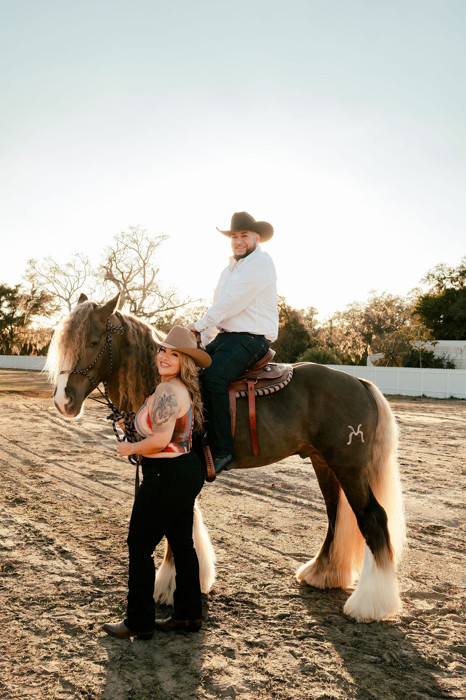 A woman stands beside a large horse while a man in a cowboy hat sits mounted in a sandy outdoor arena.