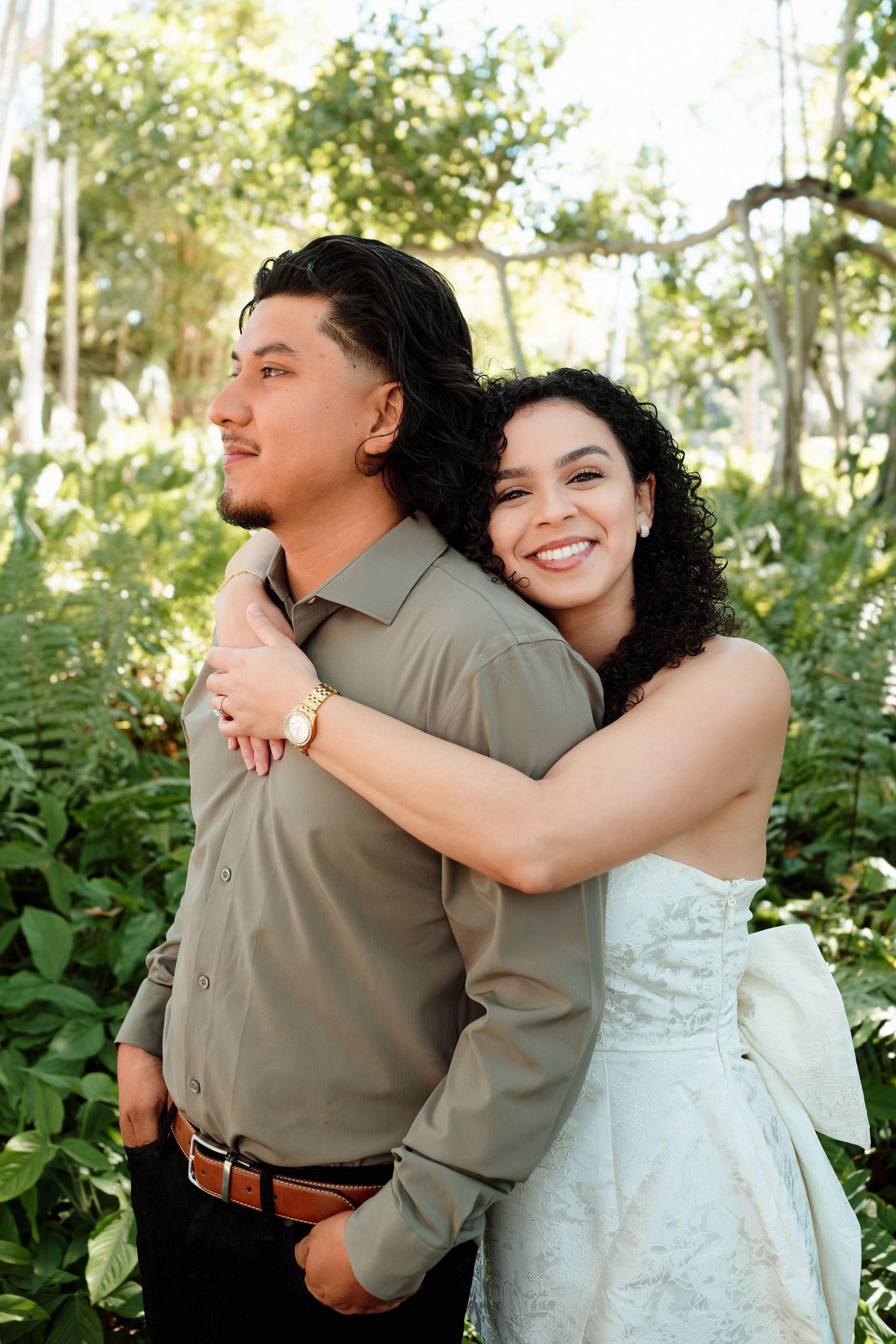 A smiling bride in a white dress embraces her groom from behind in a lush green outdoor setting.