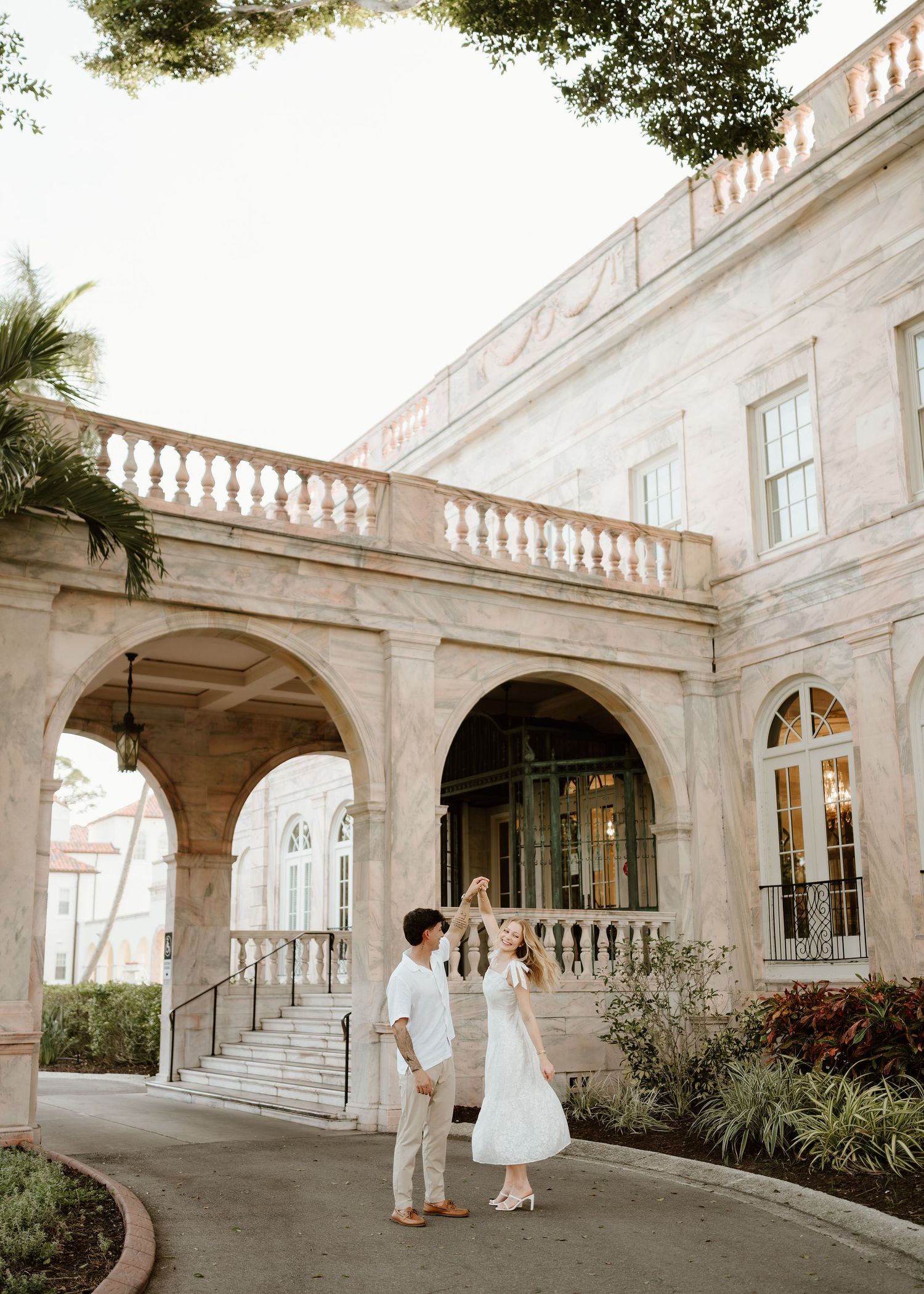 Couple dancing in white outfits at a grand Mediterranean-style estate with arched colonnades and balustrades.