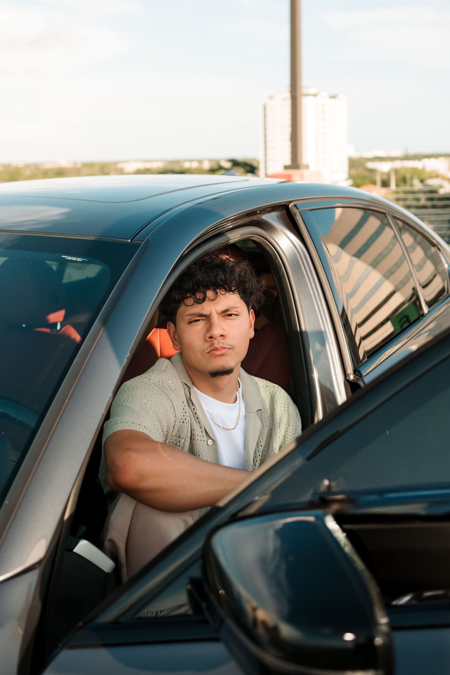 Young man with curly hair sitting in a dark car, leaning out the window, looking at the camera on a sunny day.