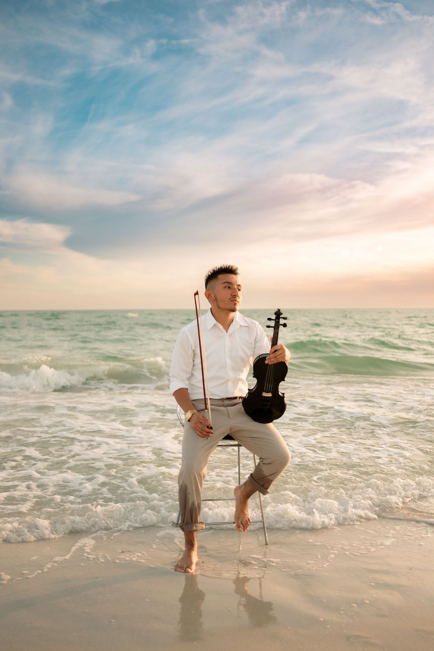 Musician holding violin and bow standing barefoot in ocean waves at sunset.