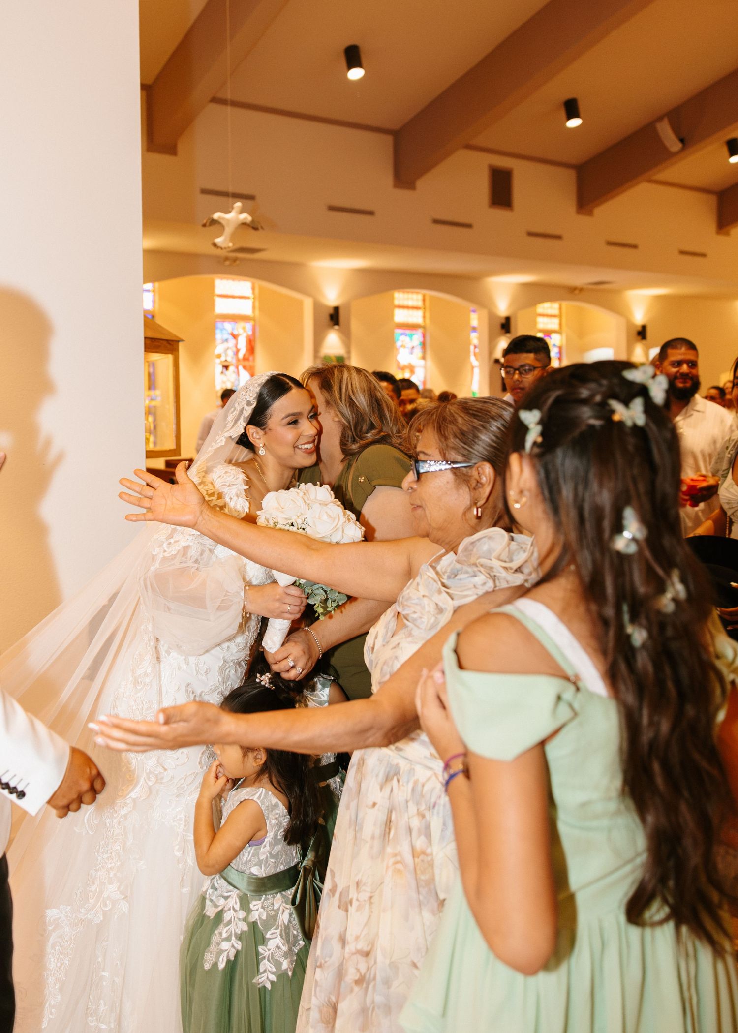 Bride being embraced by bridesmaids in sage green dresses and guests inside a warmly lit wedding chapel.