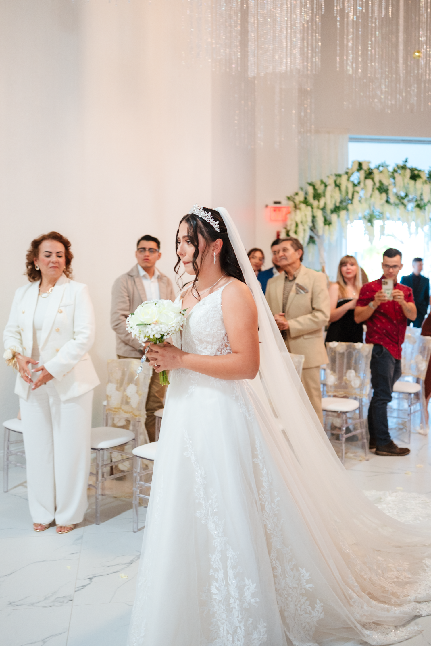 Bride in white lace gown holding bouquet walks down aisle at elegant wedding ceremony with guests seated behind her.
