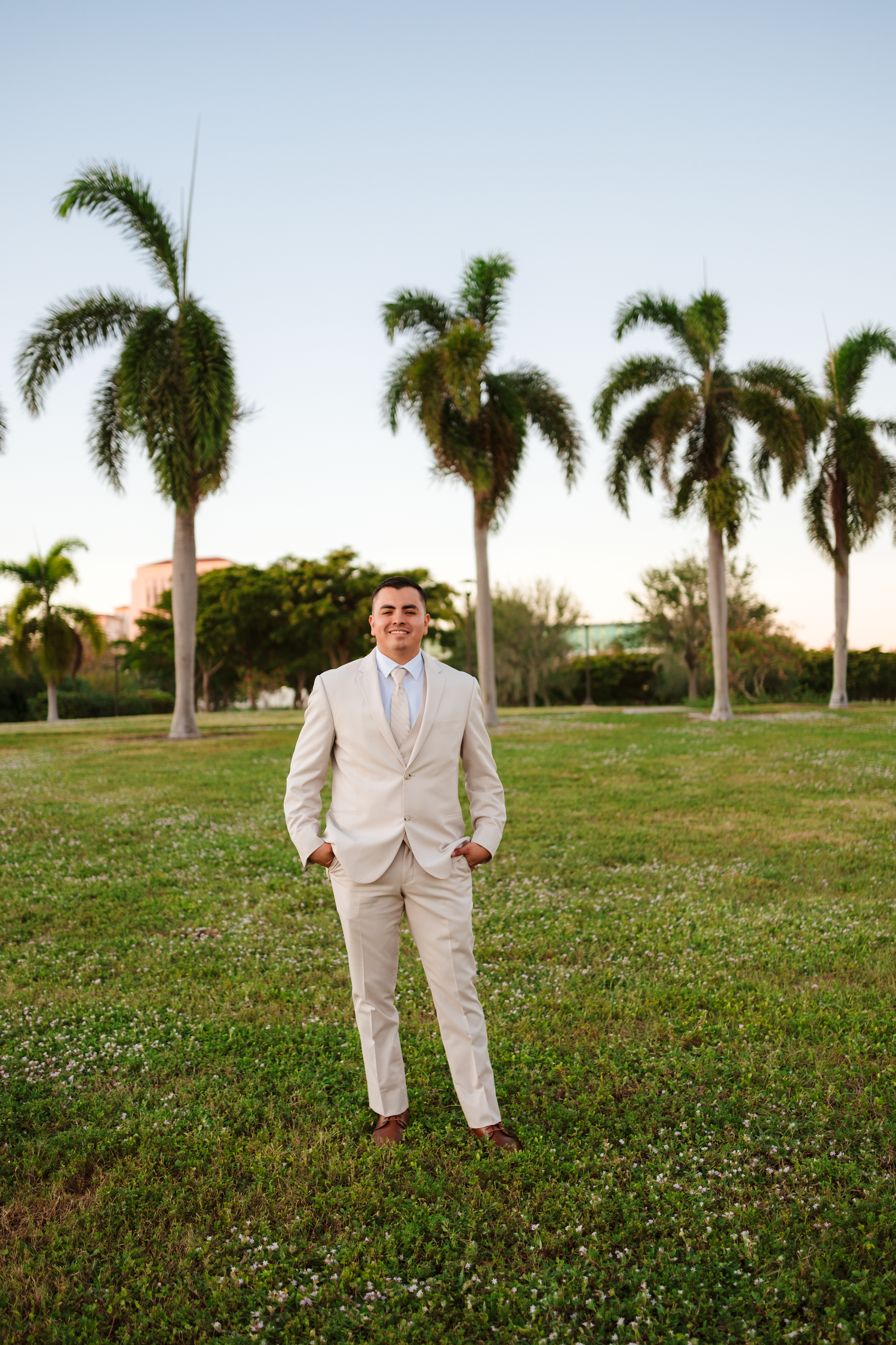 Groom in beige suit standing confidently on a lawn surrounded by tall palm trees.