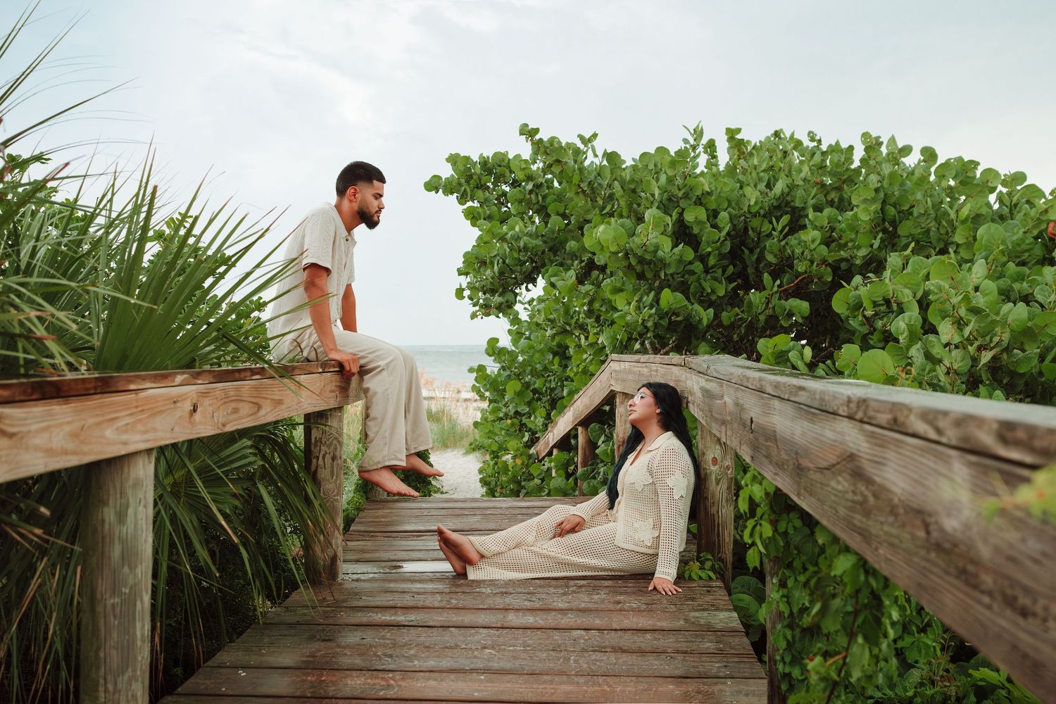 Couple on a wooden boardwalk surrounded by lush greenery near a tropical beach, one sitting on railing, one on the deck.