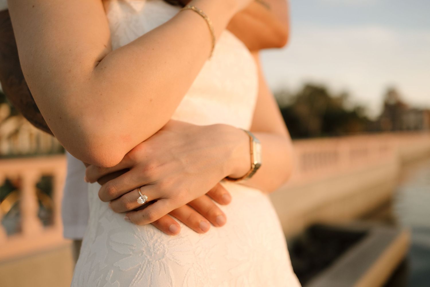Close-up of bride in white lace dress showing diamond engagement ring and gold bracelet at golden hour.