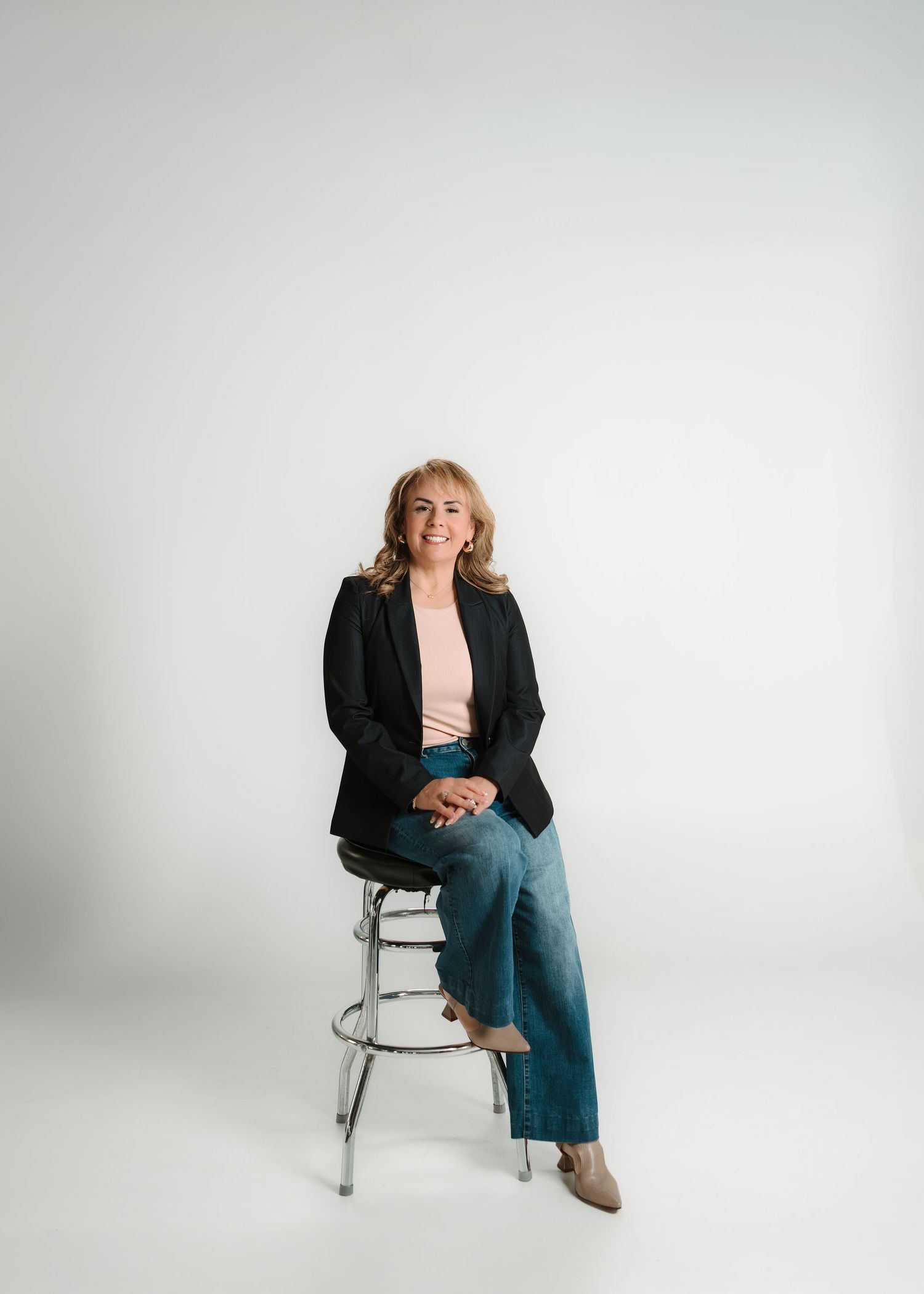 Woman in black blazer and jeans sitting on a stool against a white background, smiling.