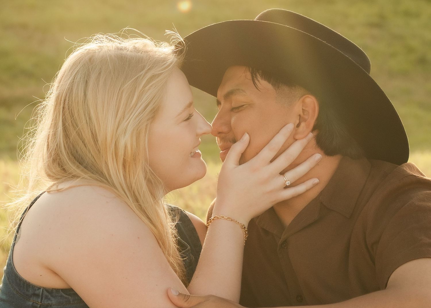 A couple shares a romantic moment outdoors, woman in blue top holding mans face as he wears a cowboy hat in golden light.