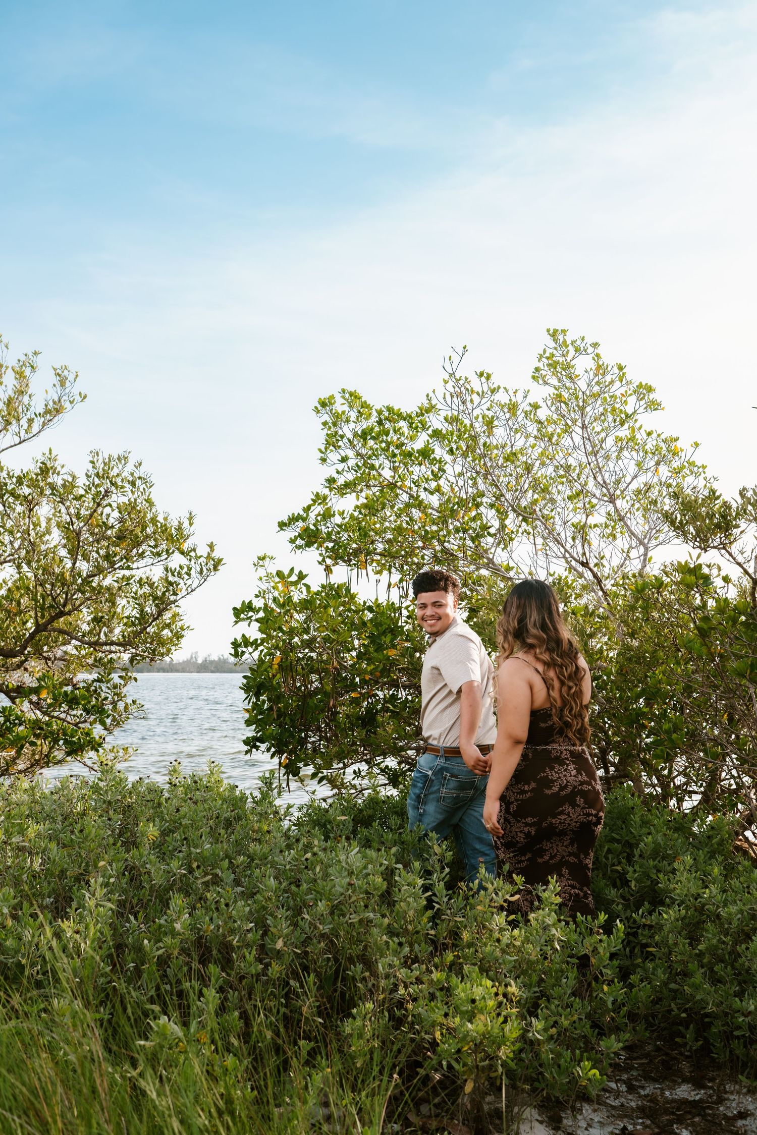 Couple holding hands among lush green trees near waterfront on a sunny day.