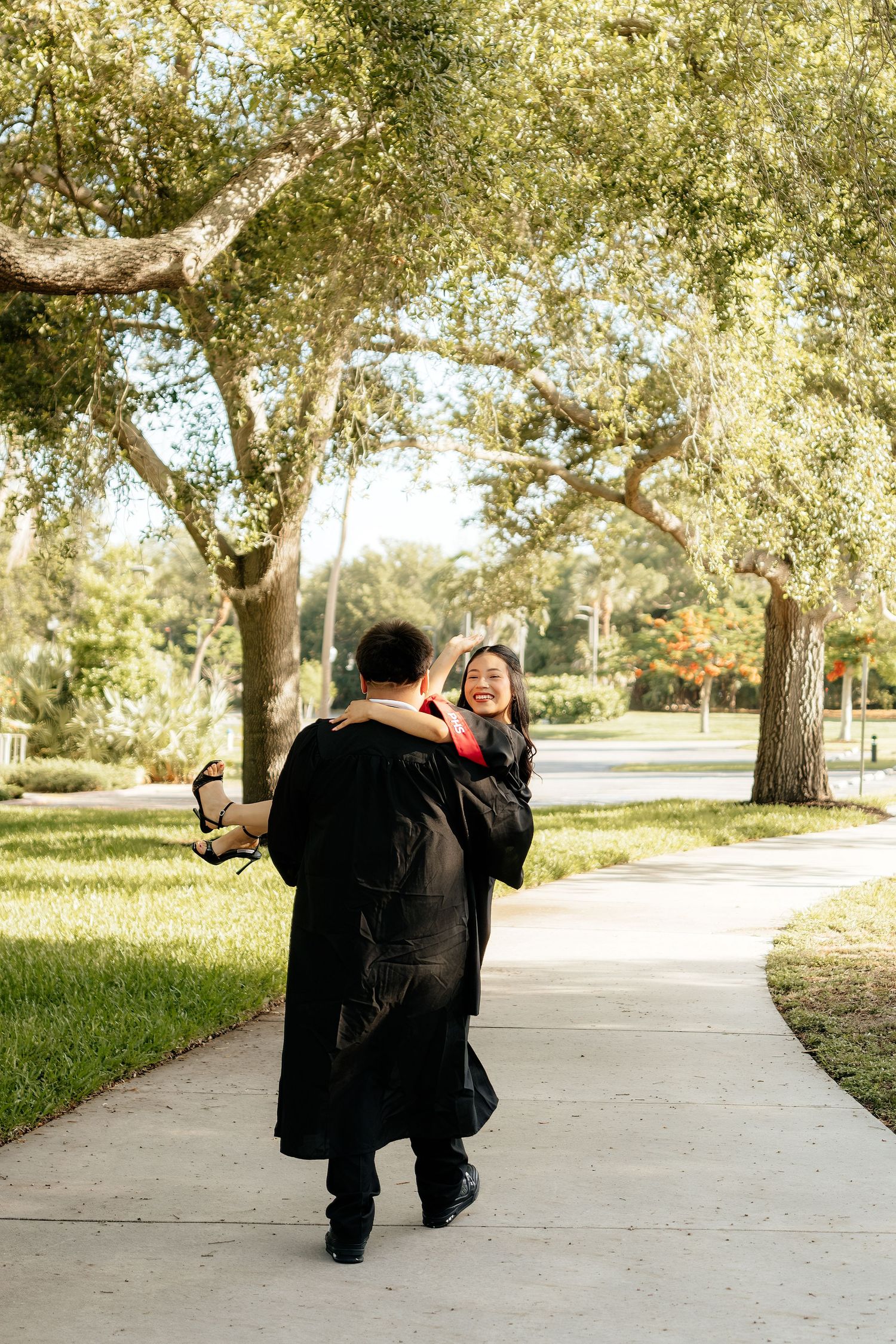 Graduate in black gown being lifted by partner on a sunny campus walkway surrounded by lush green trees.