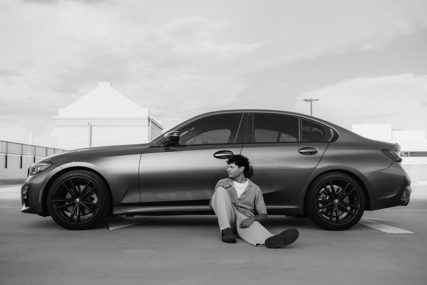 Black and white photo of a man sitting beside a sleek gray sedan in an empty parking lot.