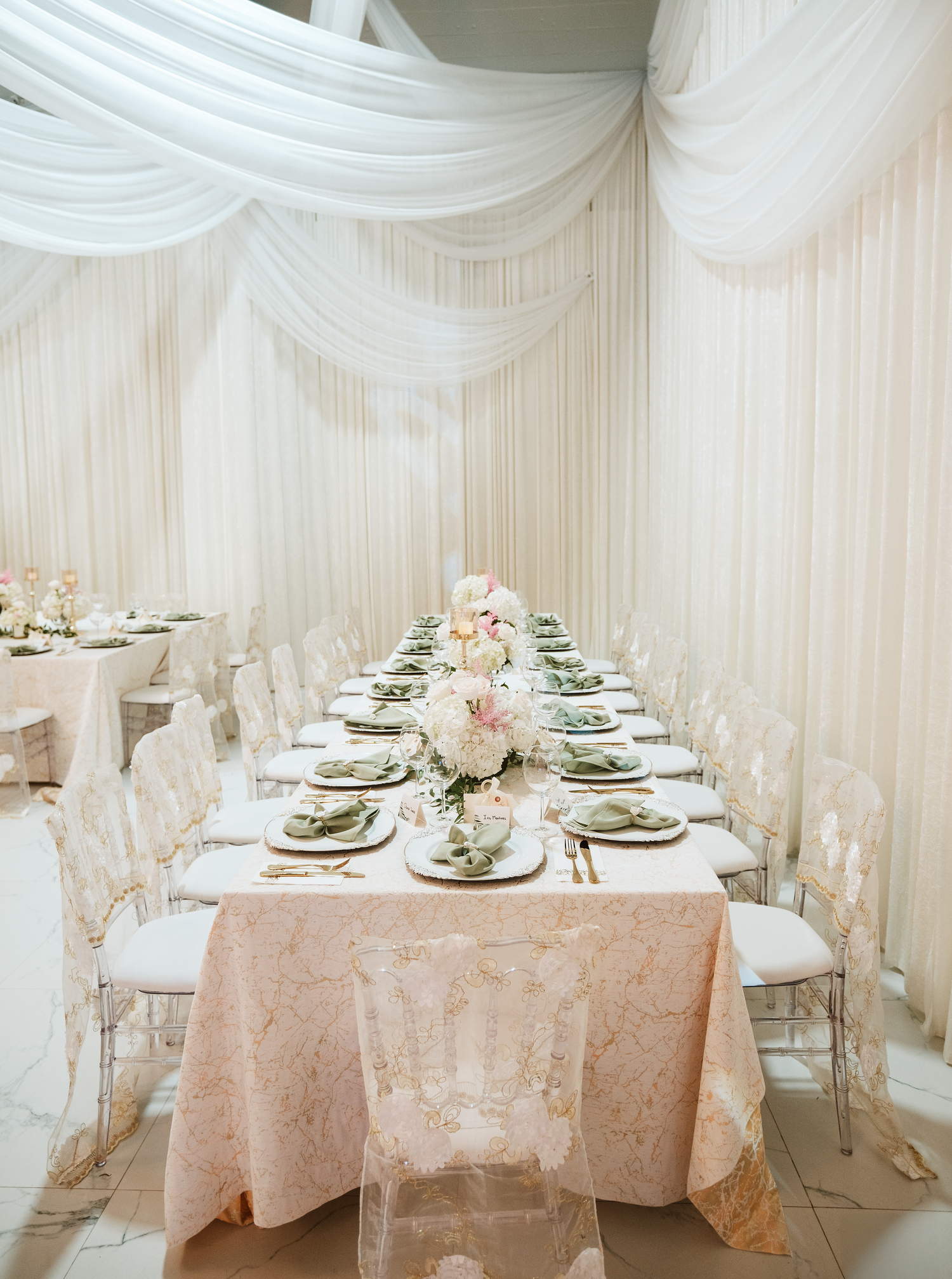 Elegant wedding reception table with blush floral linen, crystal chairs, and white draped ceiling decor.
