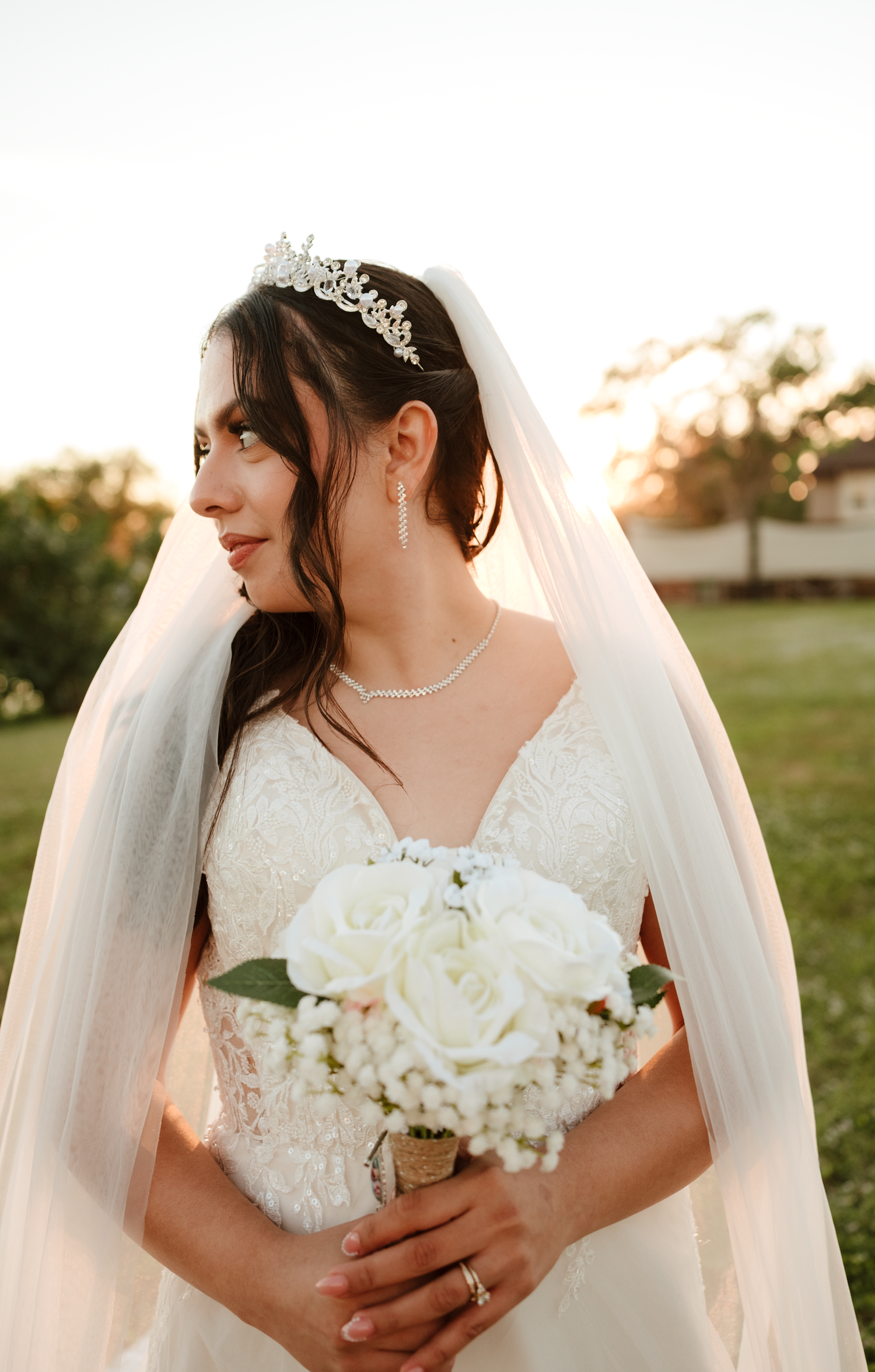 Beautiful bride wearing lace gown, crystal tiara, and veil holding white rose bouquet at sunset.