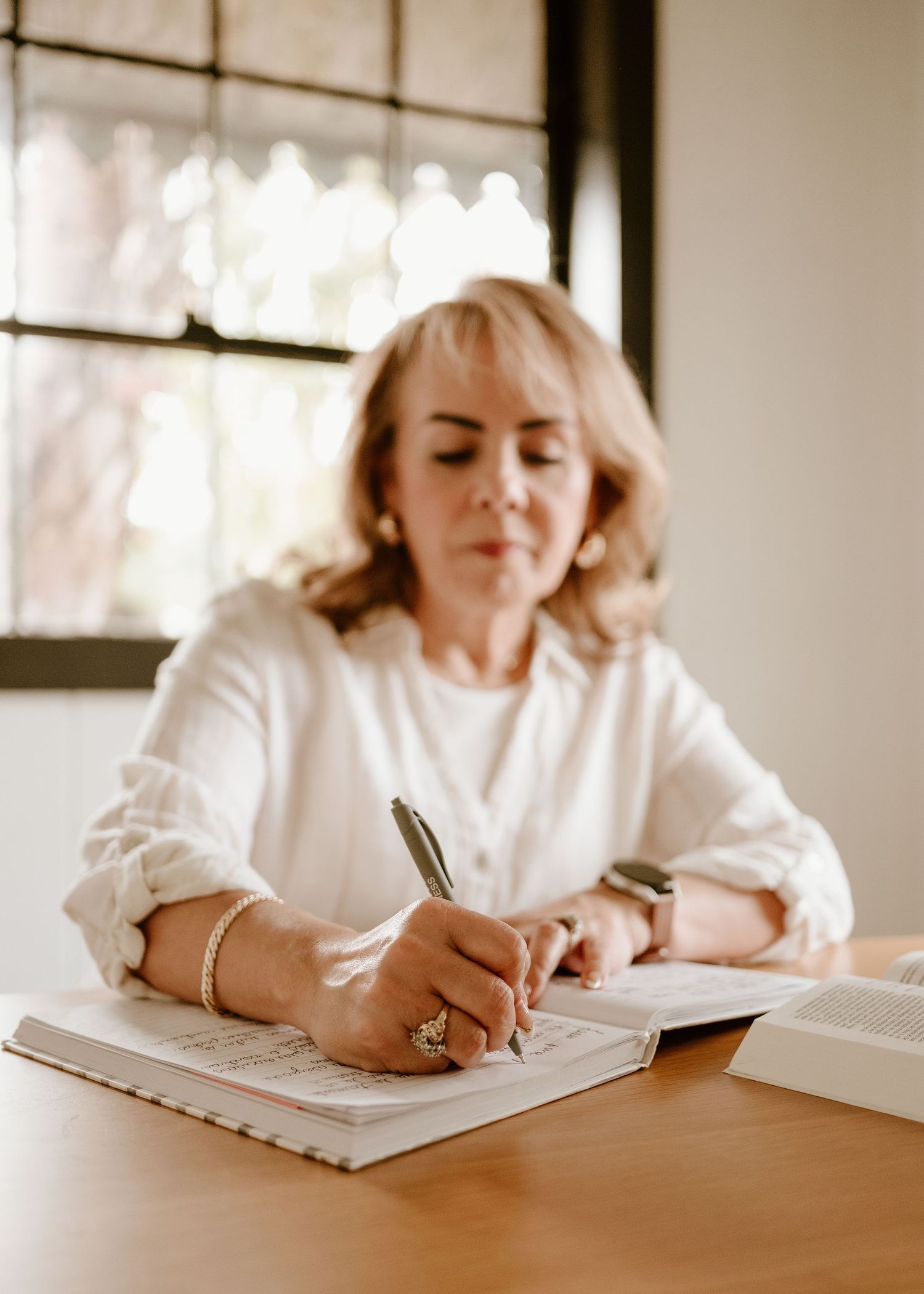 Woman in white blouse writing notes in a planner at a wooden desk with books nearby.