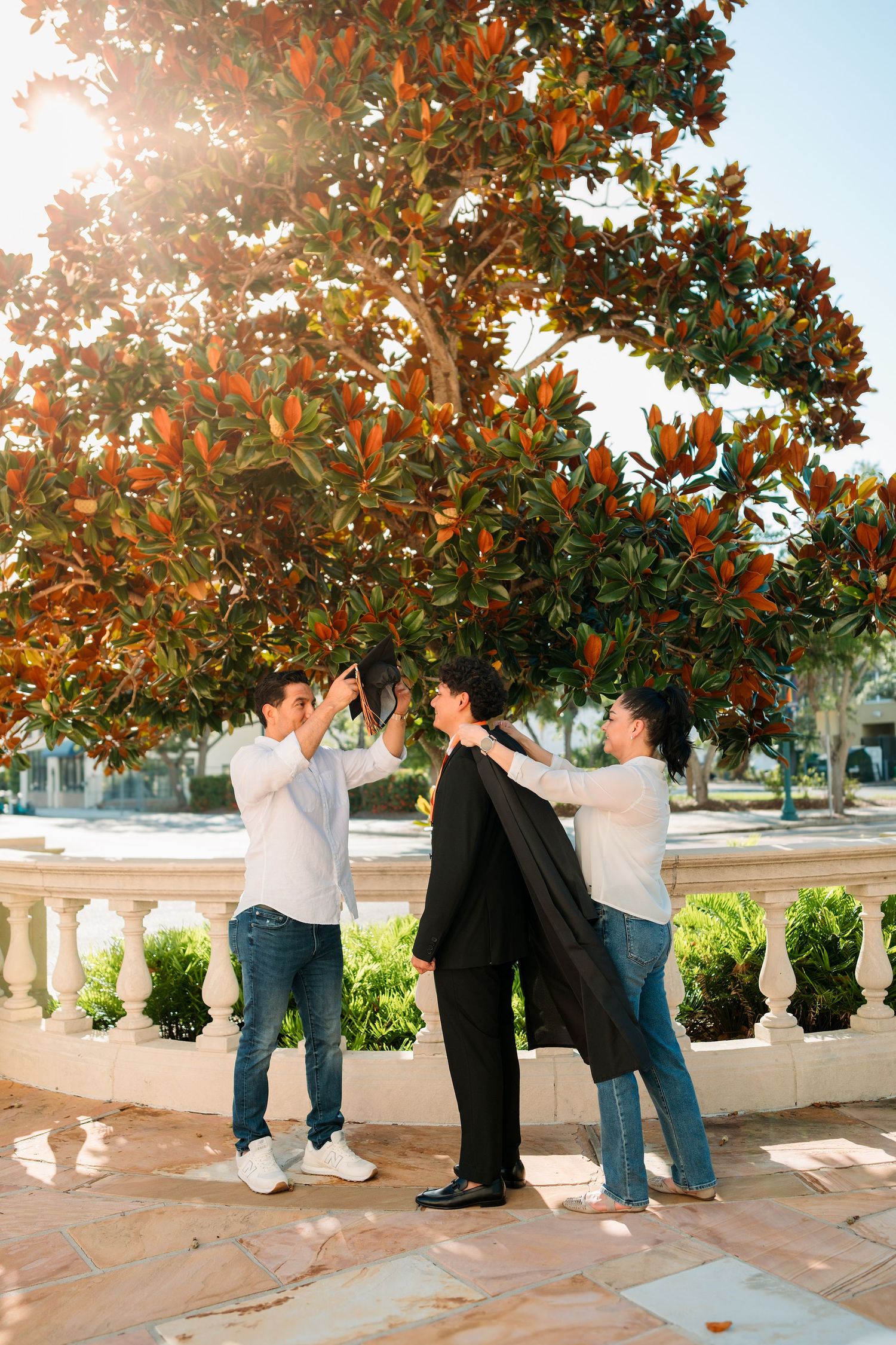 Two men help a third person put on a black suit jacket outdoors near a large tree with orange-tinted leaves.