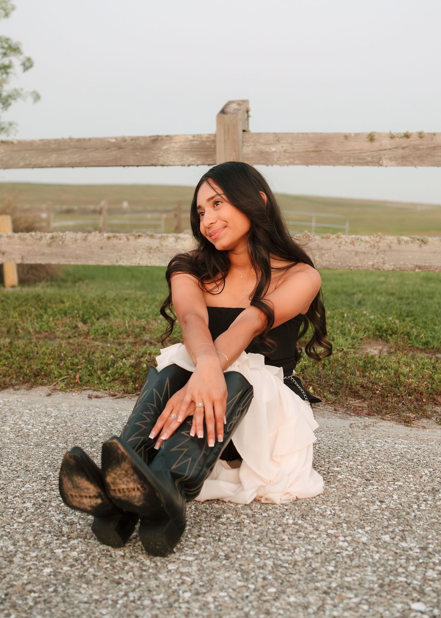 Young woman with long dark hair sitting on pavement in black top, jeans, and cowboy boots near a wooden fence.