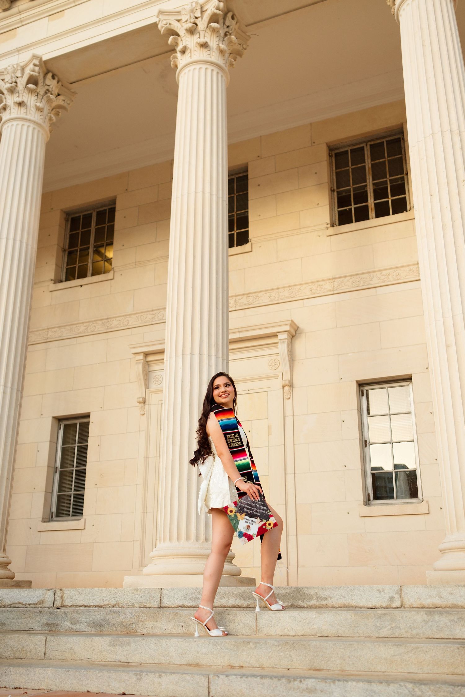 Young woman in white outfit with colorful sash poses on steps of neoclassical building with tall columns.